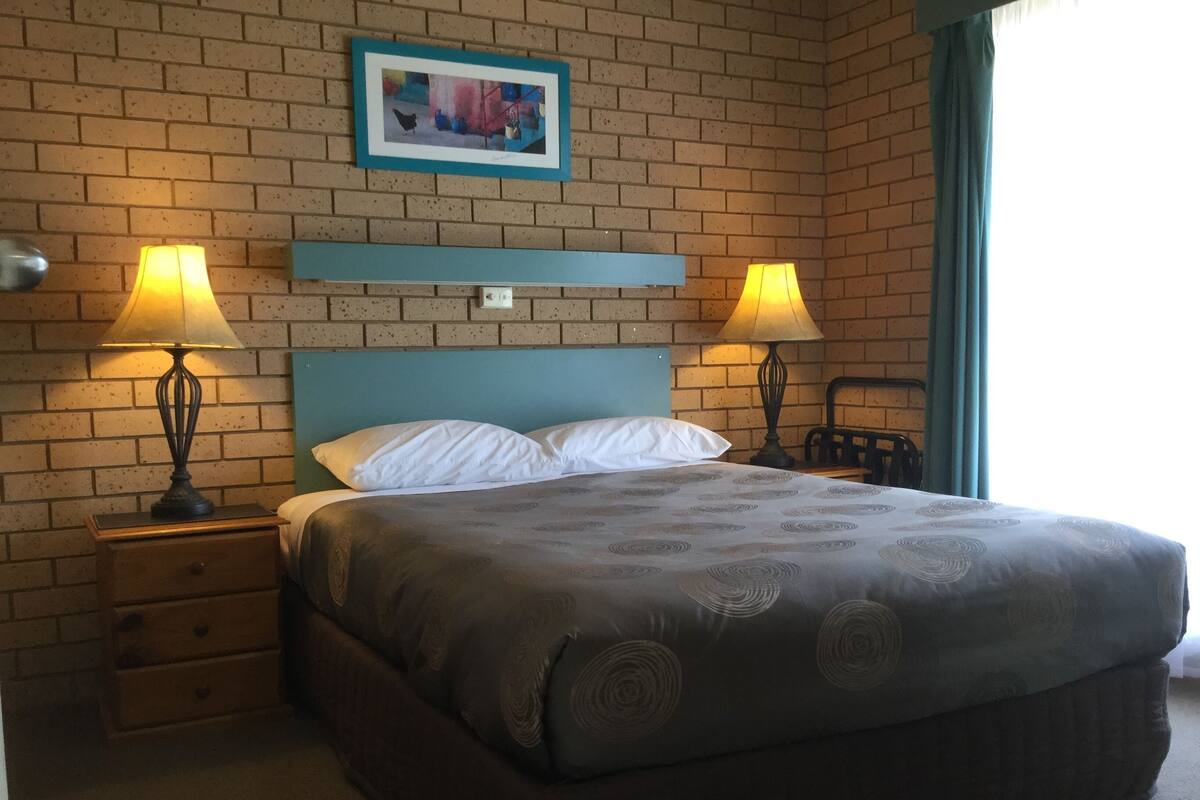 A hotel room with a brick-patterned wall, a blue headboard, a bed with a patterned bedspread, two bedside lamps, a small wooden nightstand with drawers, and a framed picture above the headboard.