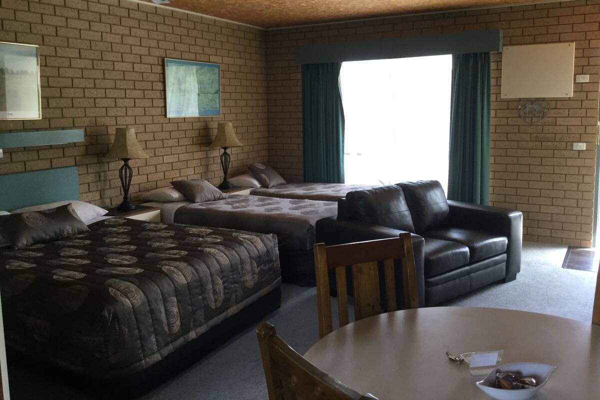 A hotel room with a brick wall, two beds, a leather sofa, a wooden table, and a ceiling fan.