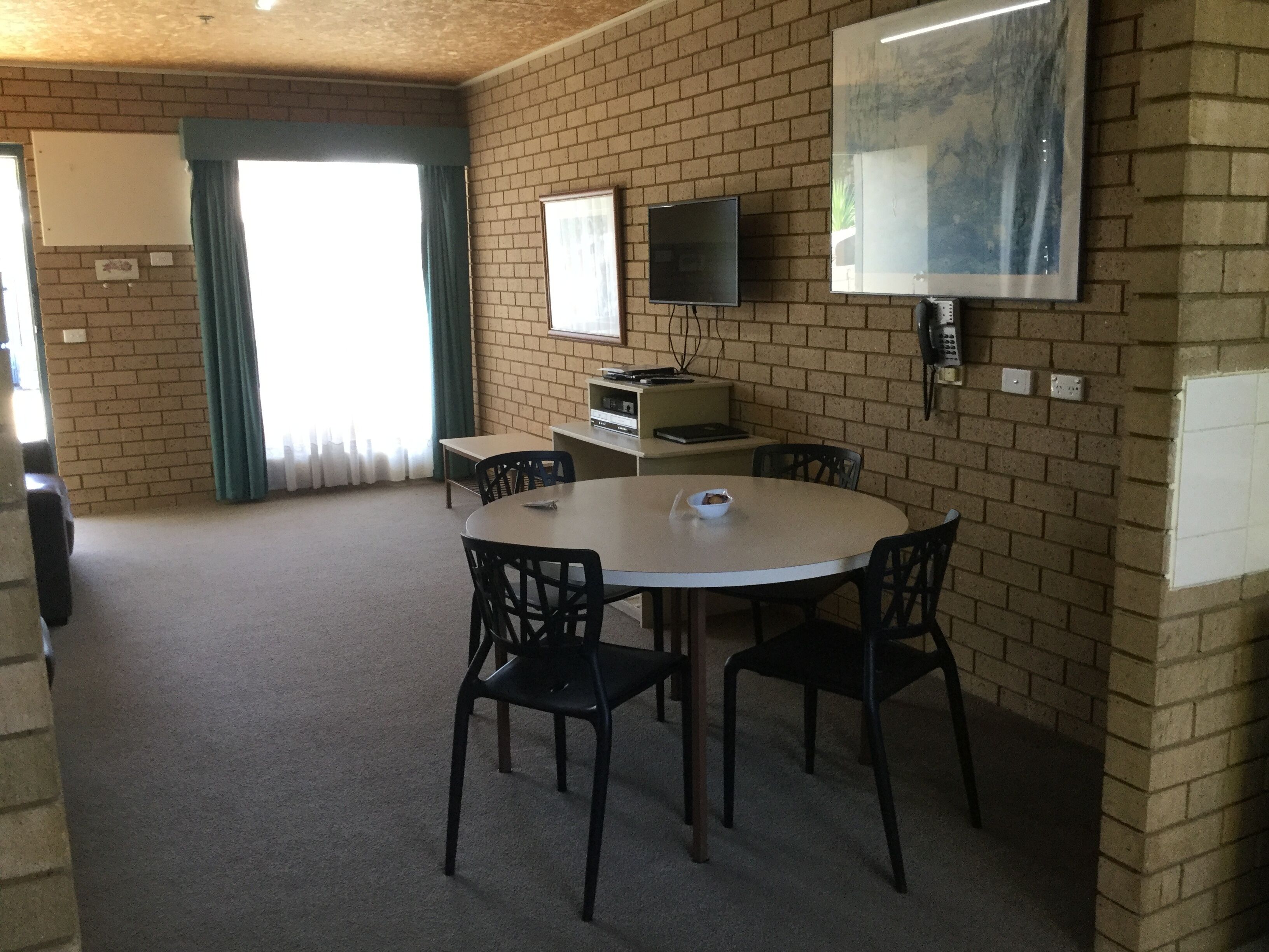 A room with a brick wall, a round table with four chairs, a flat-screen TV mounted on the wall, and a window with curtains.