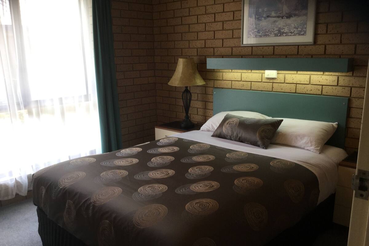 A hotel room with a brick wall, a bed with a patterned bedspread, a nightstand with a lamp, and a framed picture above the bed.