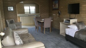 A compact living space with a sofa, a dining table with chairs, a small TV, and a kitchen area with a window. - Sunrise Motel (Barooga)