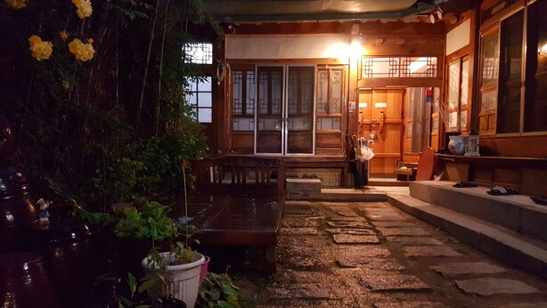 Um edifício japonês tradicional com fachada de madeira, um caminho de pedra que leva à entrada e plantas em vasos.
