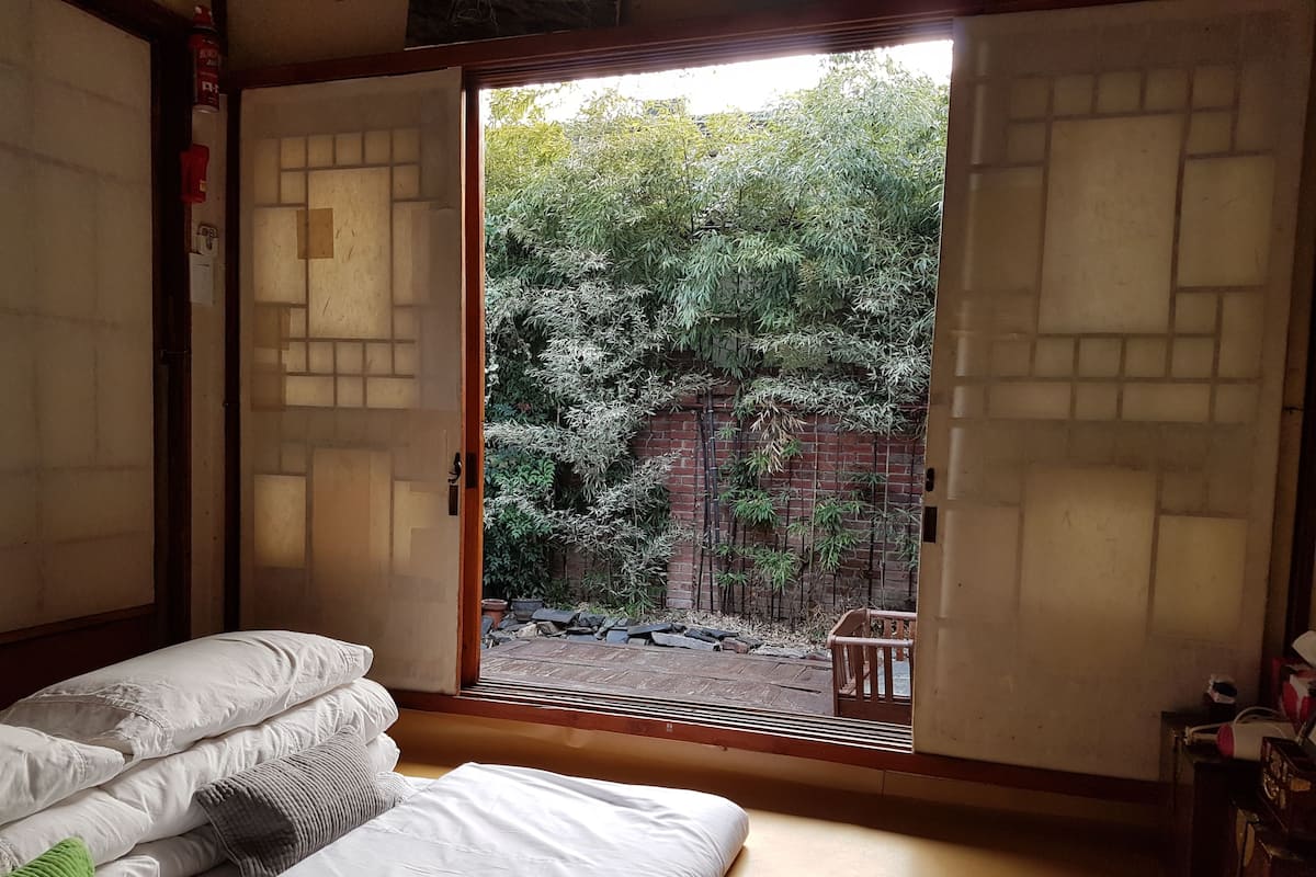 A room with a bed, pillows, and a sliding door leading to an outdoor area with bamboo plants and a brick wall.