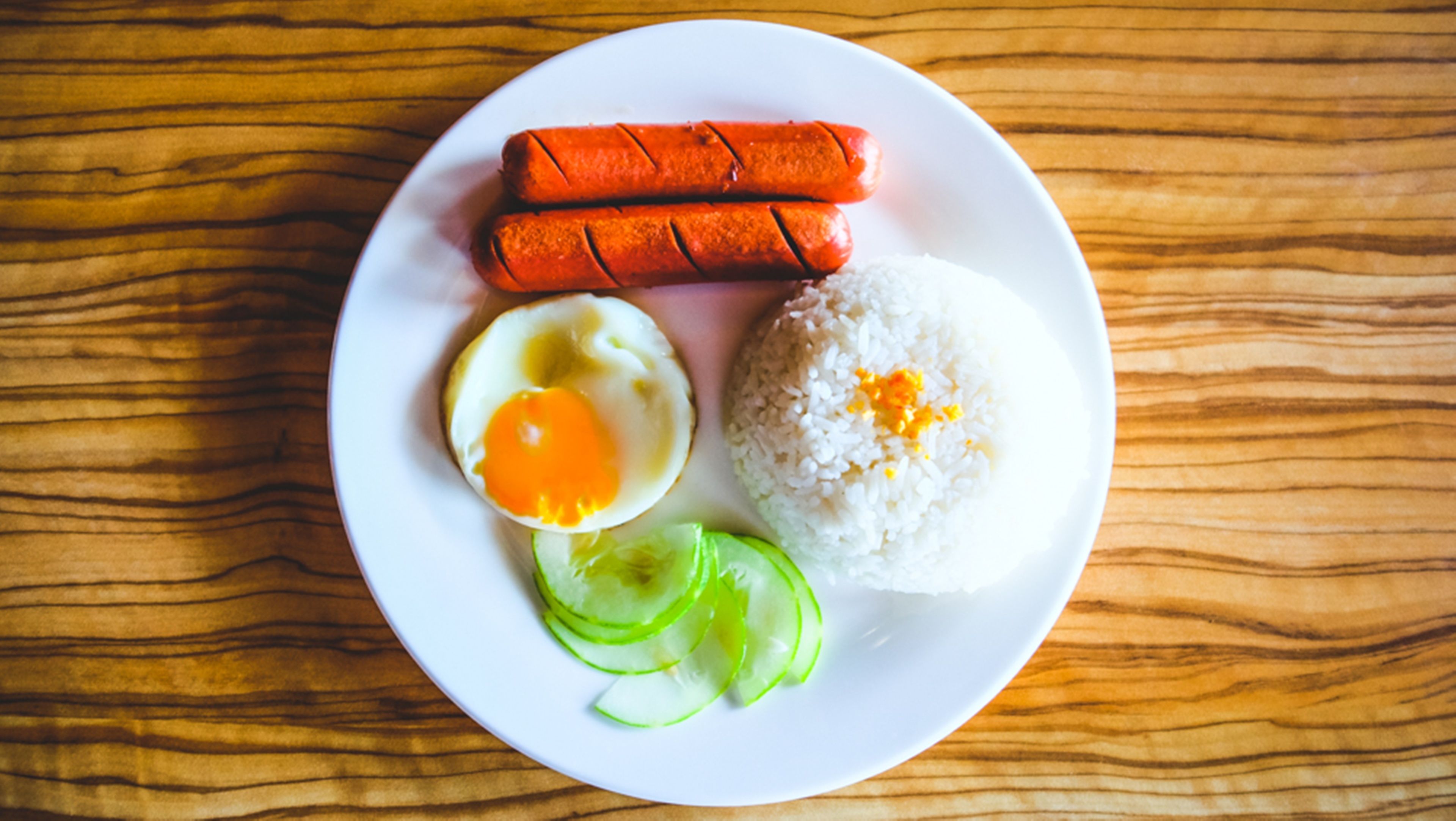 A plate with two sausages, a fried egg, rice, and cucumber slices on a wooden table.