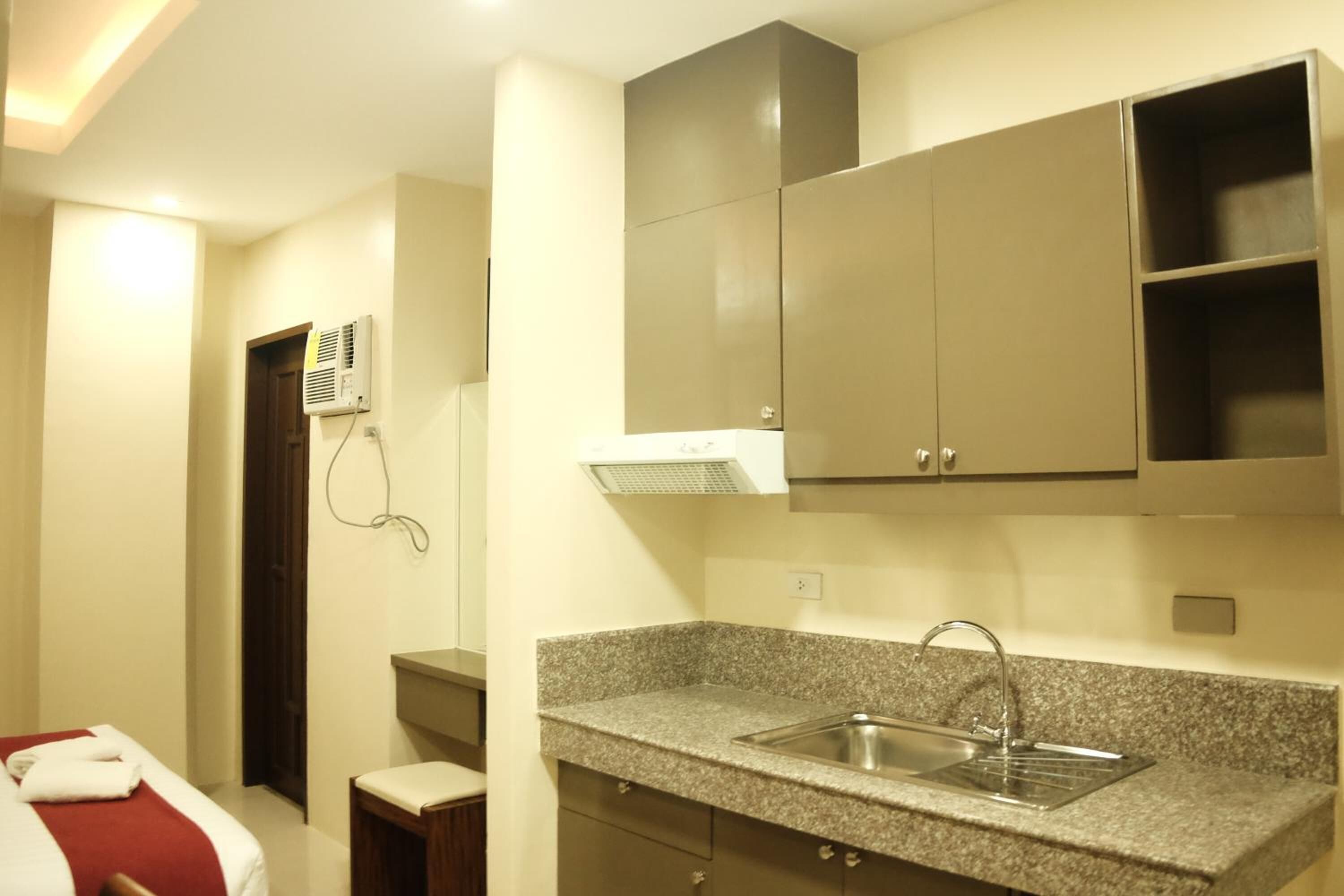 A compact hotel room with a kitchenette, a bed with a red and white bedspread, and a small wooden stool.