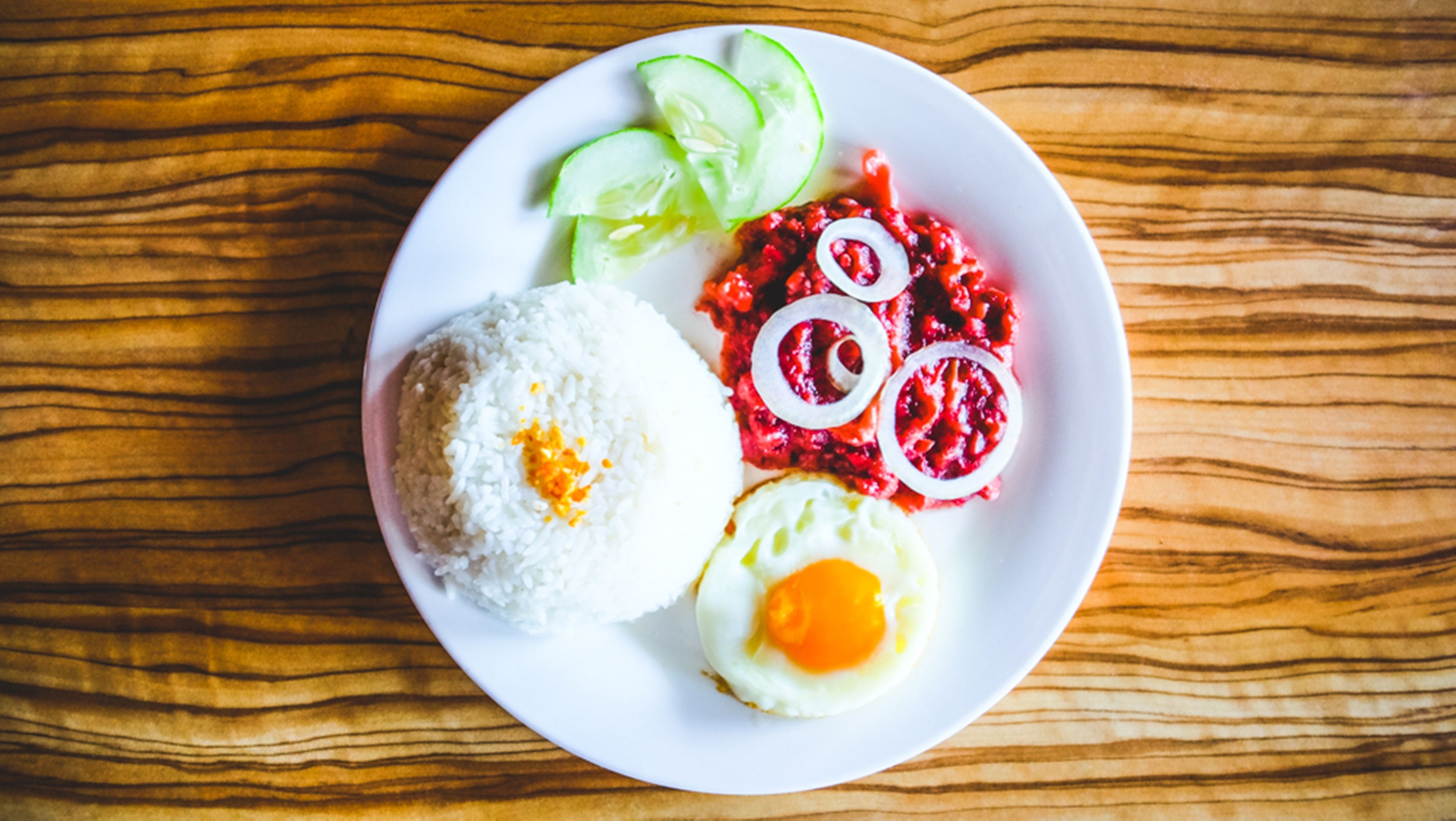 Un plato con arroz blanco, un huevo frito, verduras encurtidas rojas y rodajas de pepino sobre una mesa de madera.