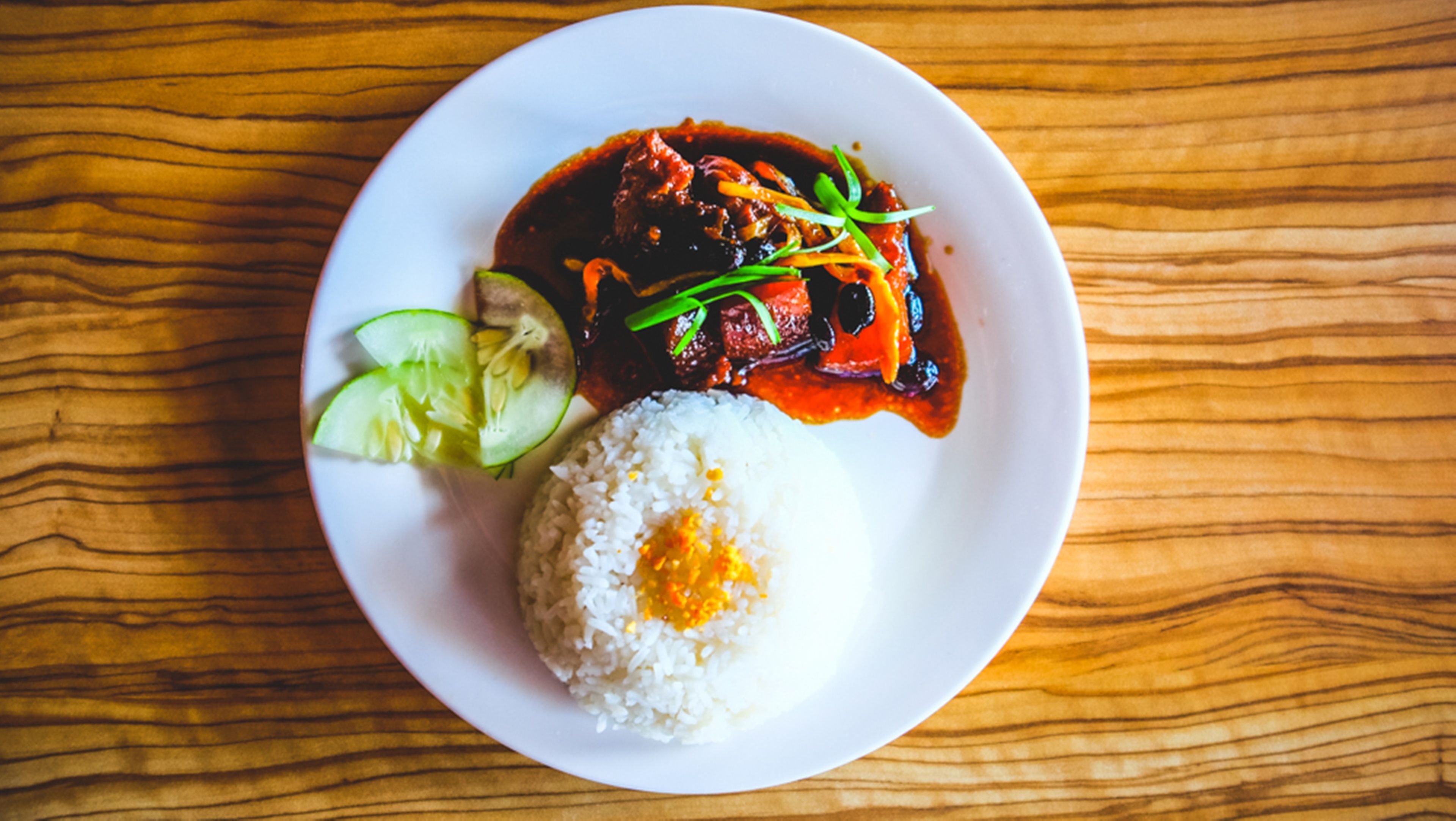 A plate of food with rice, a meat dish, and sliced cucumbers on a wooden table.