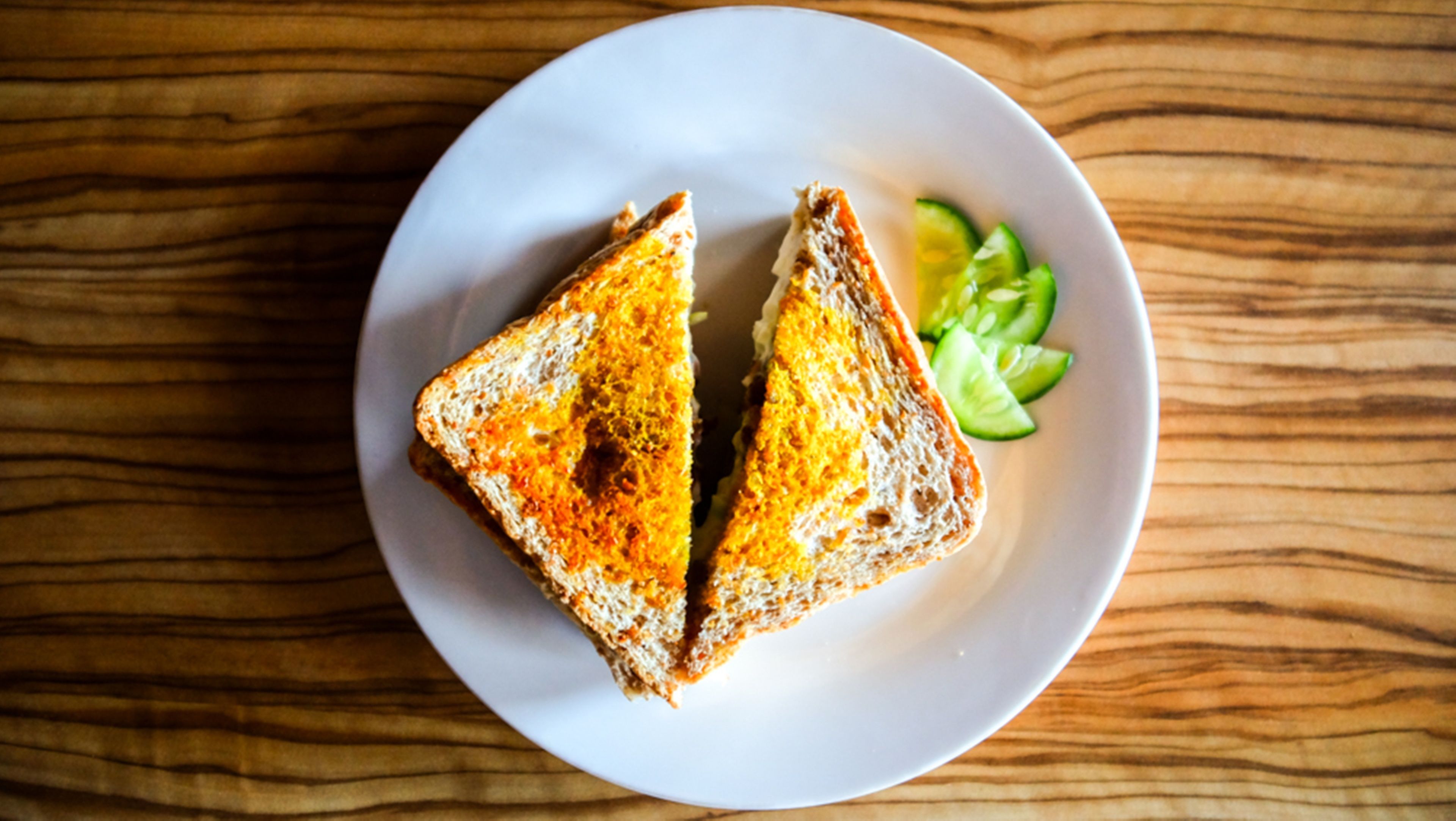 A sandwich on a white plate with cucumber slices on a wooden table.