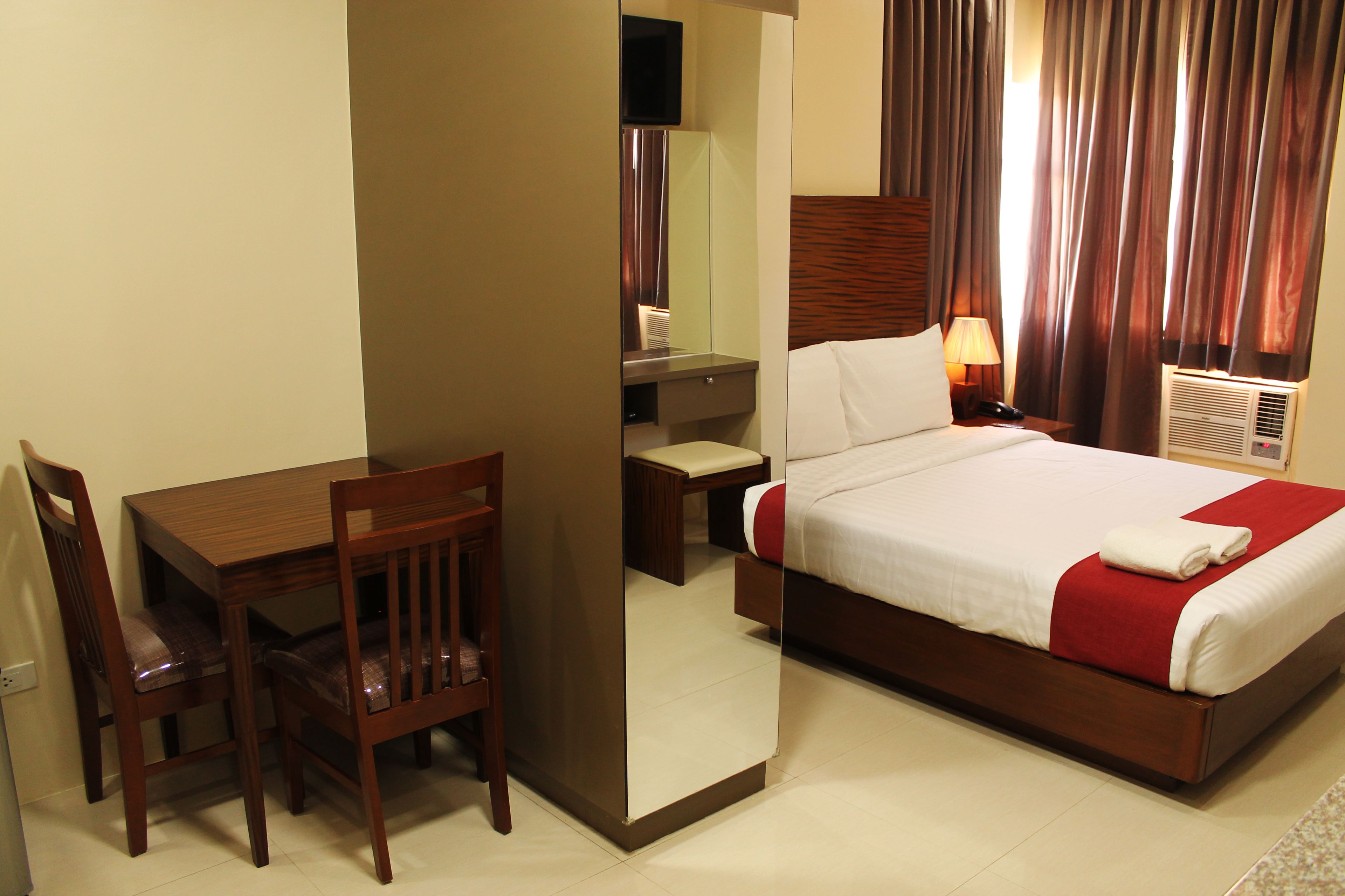 A hotel room with a bed, a desk, a chair, a mirror, a nightstand, and a lamp.