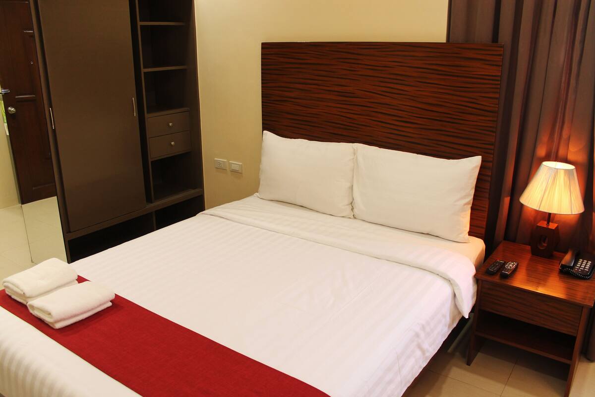 A neatly made bed with white linens and a red accent, a wooden headboard, a bedside table with a lamp and remote controls, and a wardrobe with a mirror and a hanging rack.
