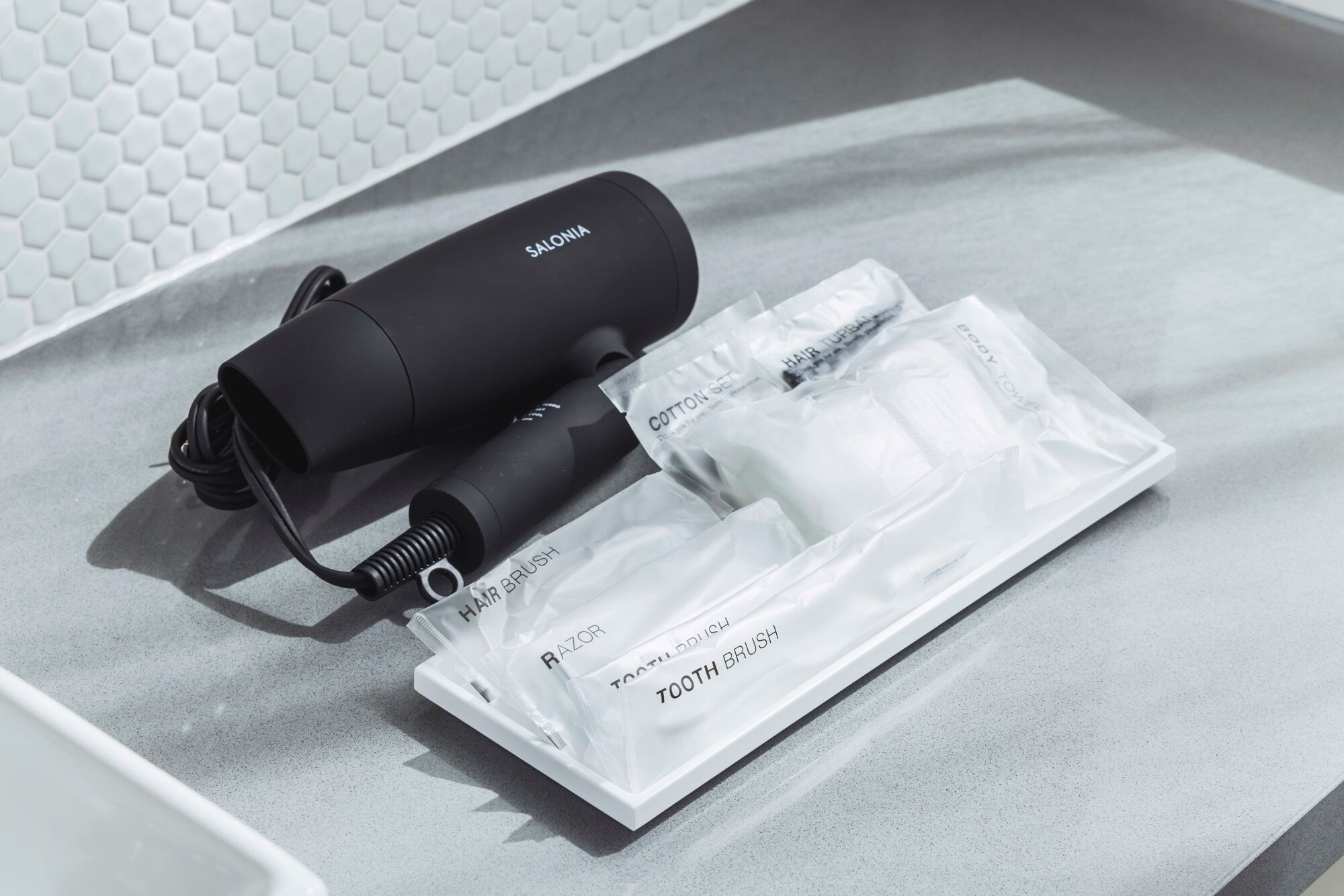A black hair dryer with a cord, a white cotton set, and a razor brush on a grey surface.