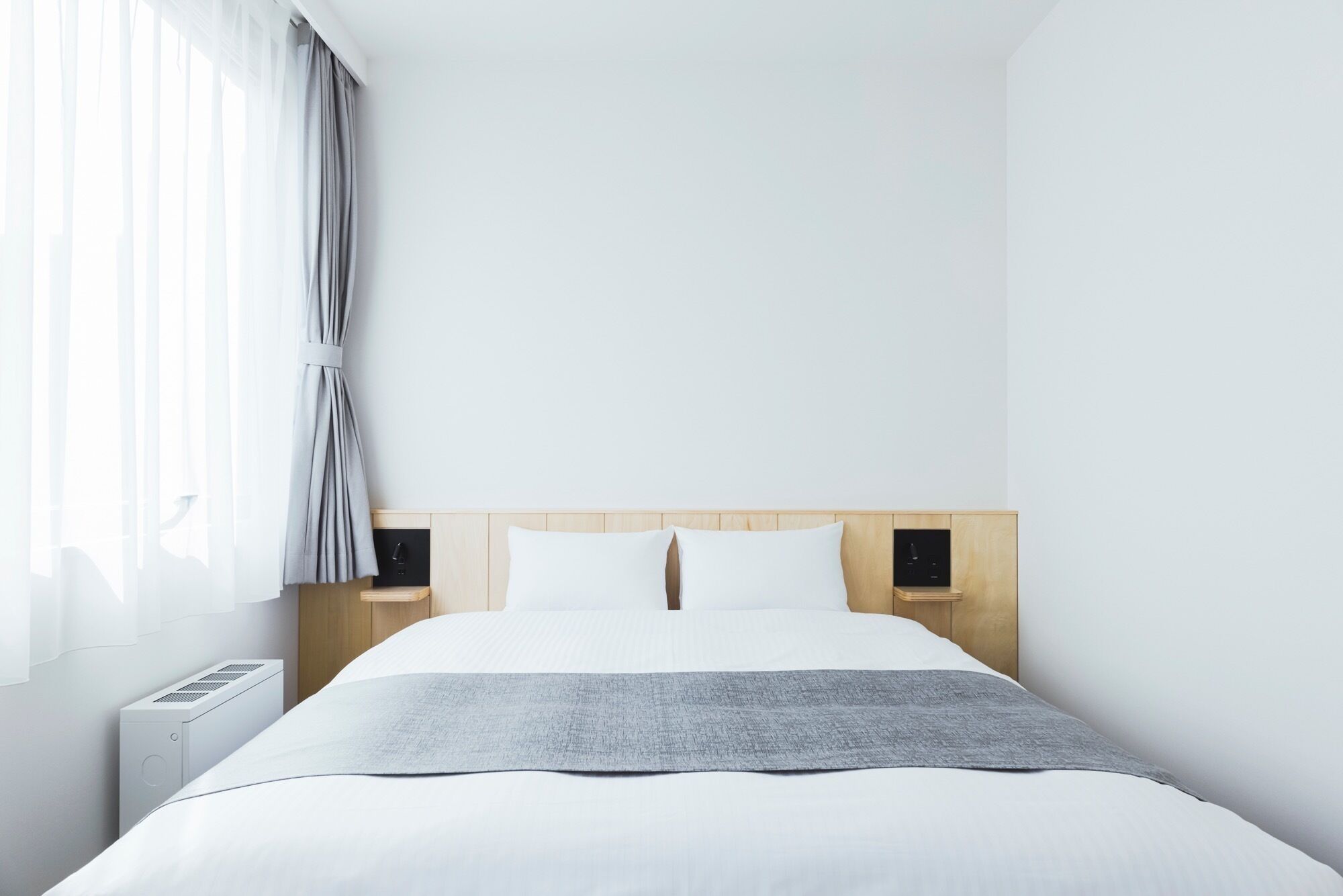 A minimalist hotel room with a large bed, a bedside table, a nightstand, and a window with curtains.