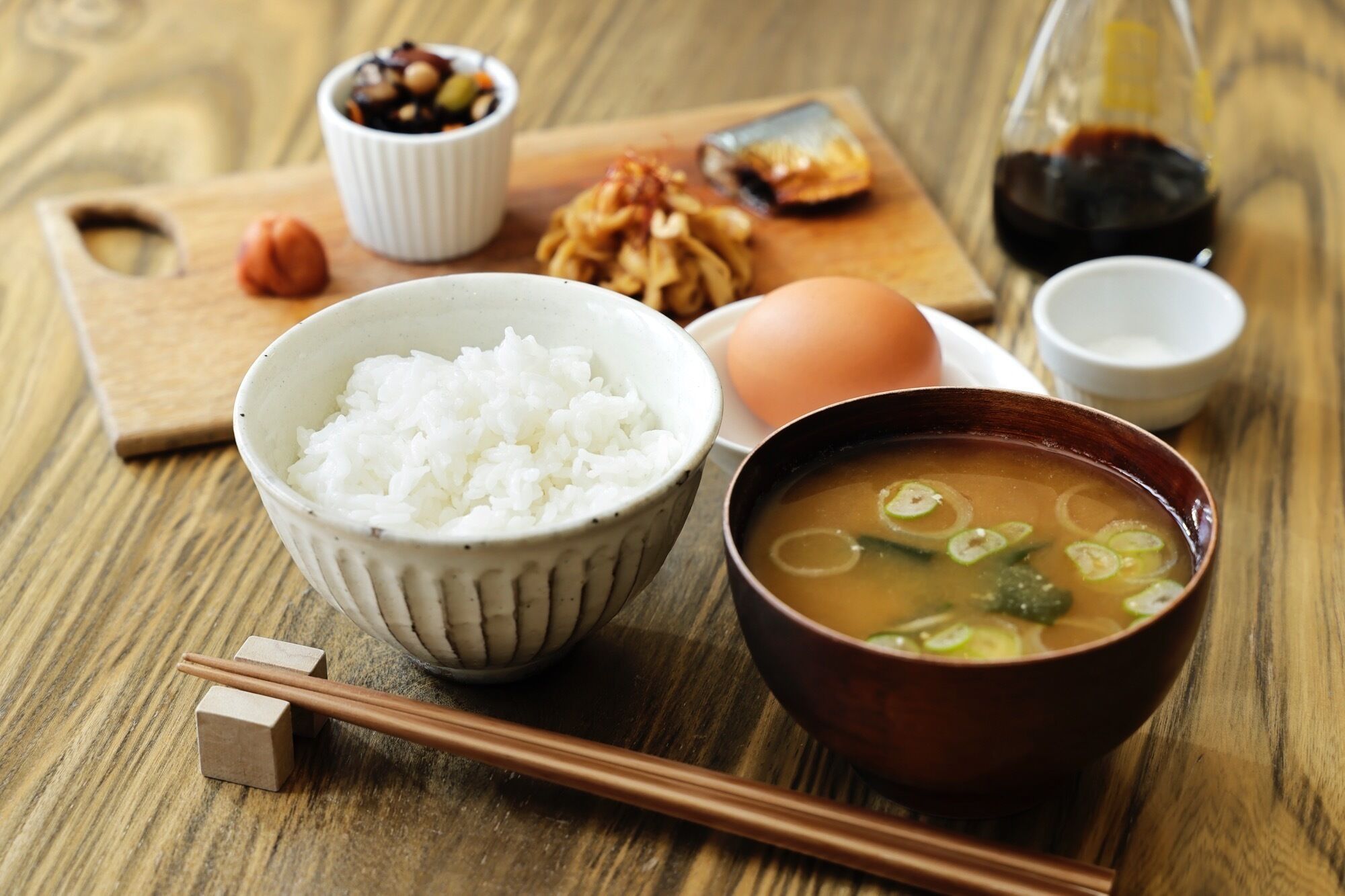 Uma mesa de madeira com uma tigela de arroz branco, uma sopa com cebolinha, um ovo inteiro, uma pequena tigela de castanhas e um frasco de molho de soja, além de hashis.