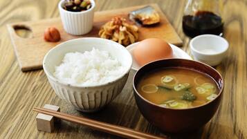 Uma mesa de madeira com uma tigela de arroz branco, uma sopa com cebolinha, um ovo inteiro, uma pequena tigela de castanhas e um frasco de molho de soja, além de hashis.
