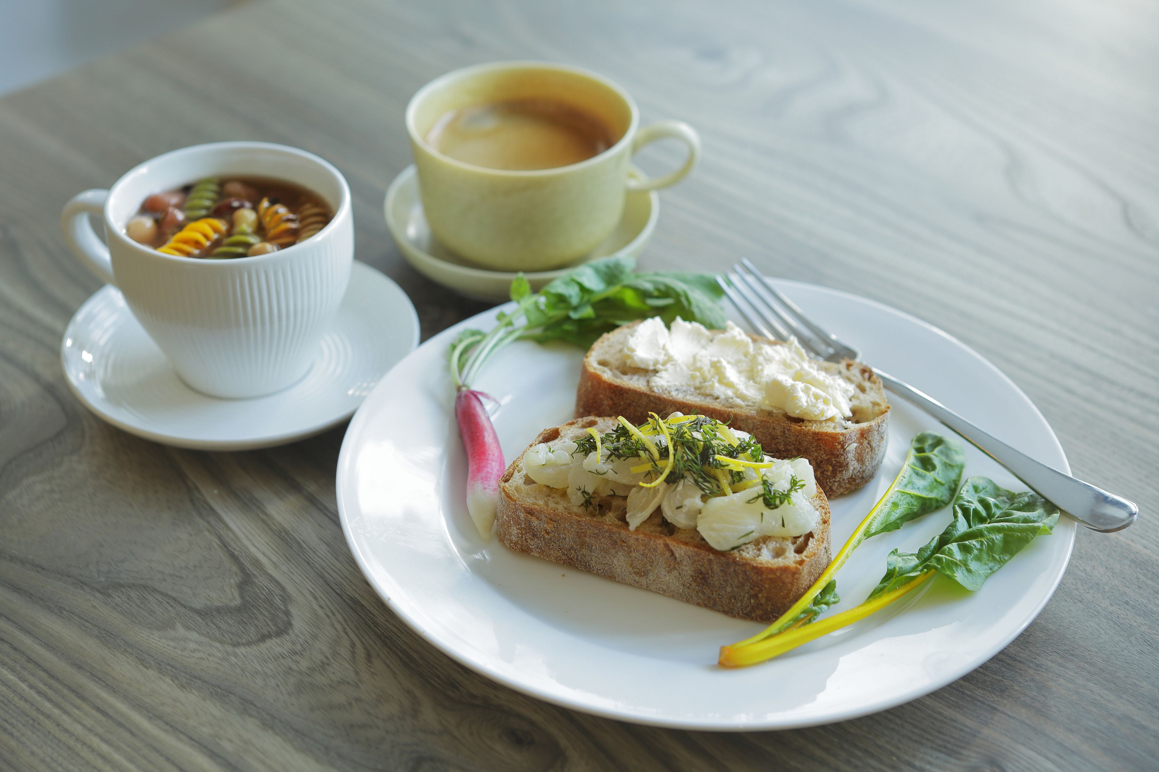 A plate with two slices of bread topped with a white spread and greens, a cup of tea, and a small bowl of soup.