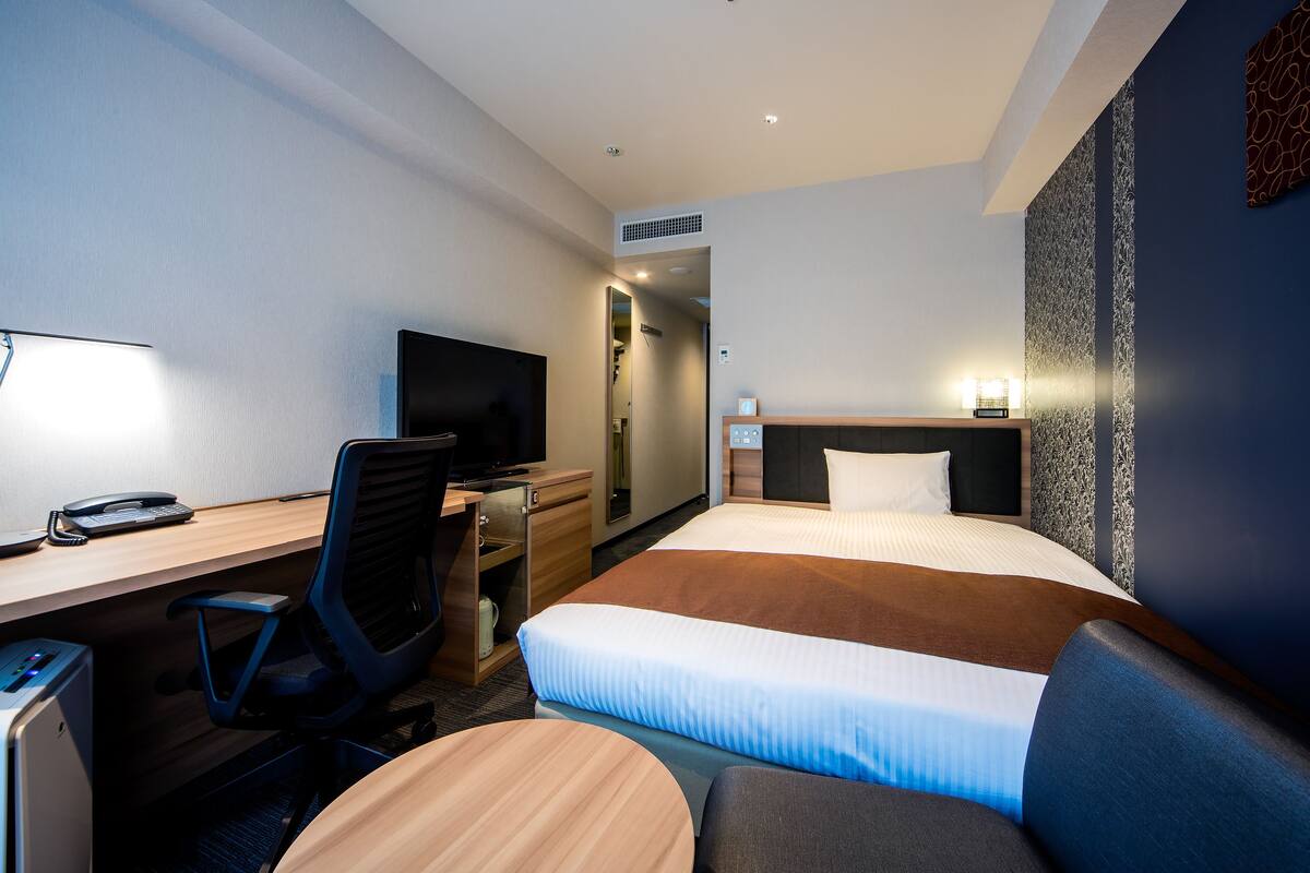 A hotel room with a bed, desk, chair, TV, and a small table.