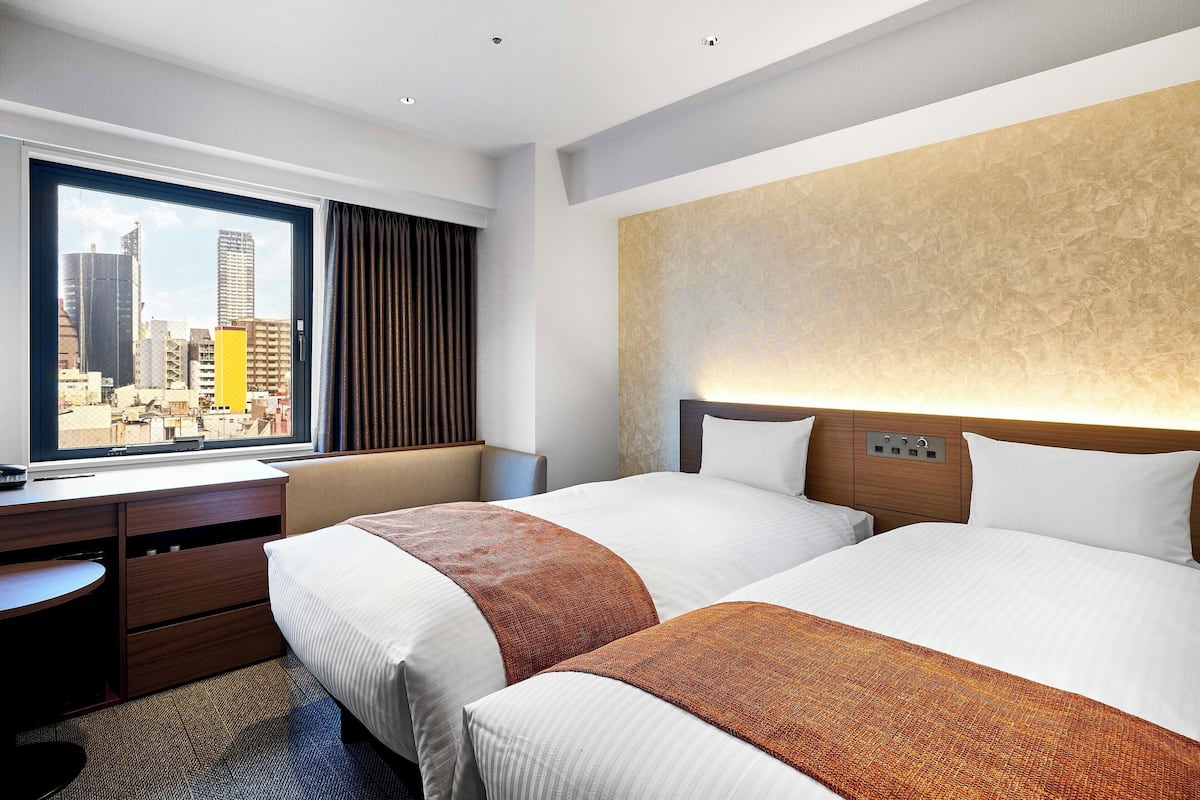 A hotel room with two beds, a desk, and a window with a city view.