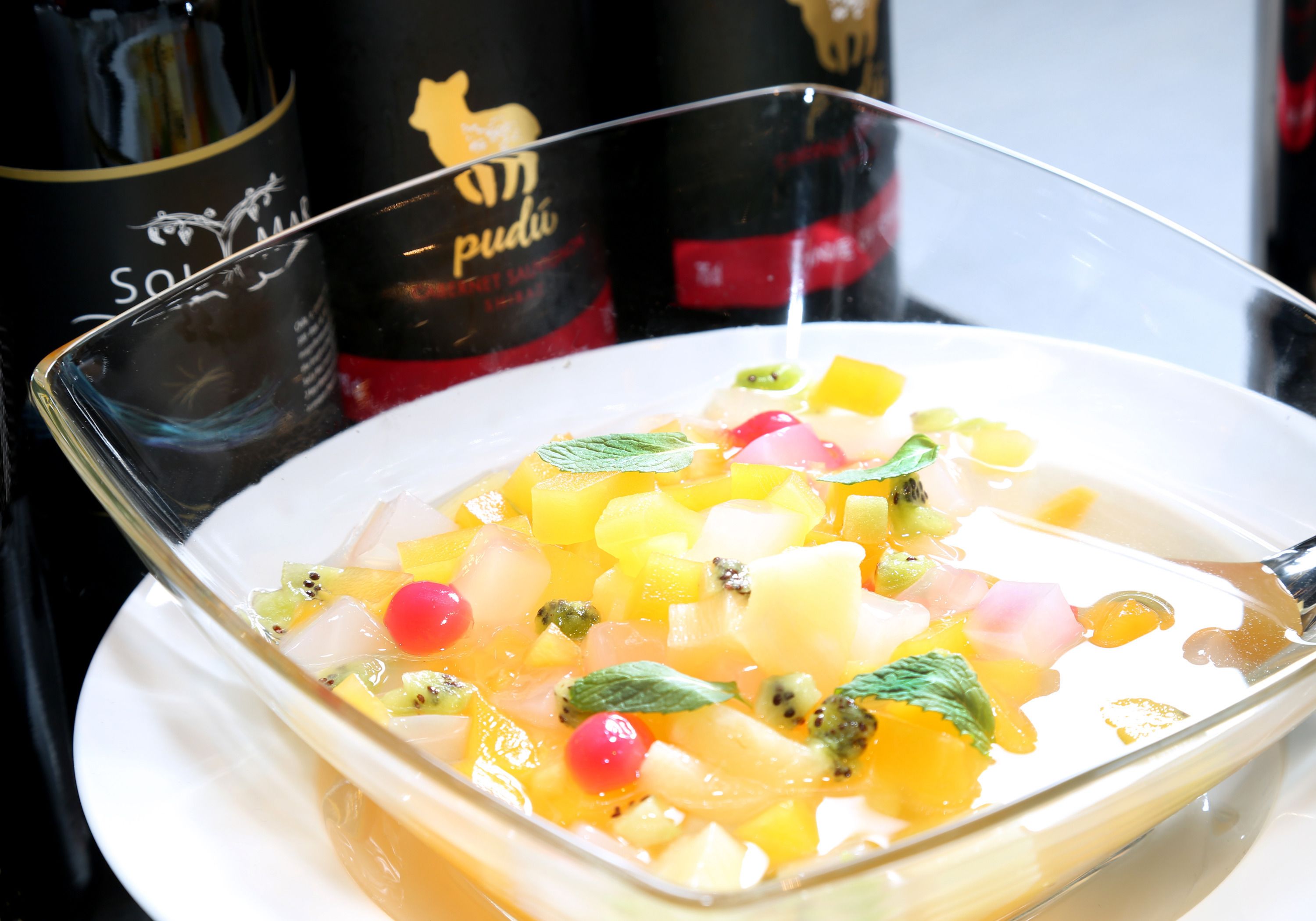 A dessert dish with fruit and mint, a glass, and two bottles of wine in the background.