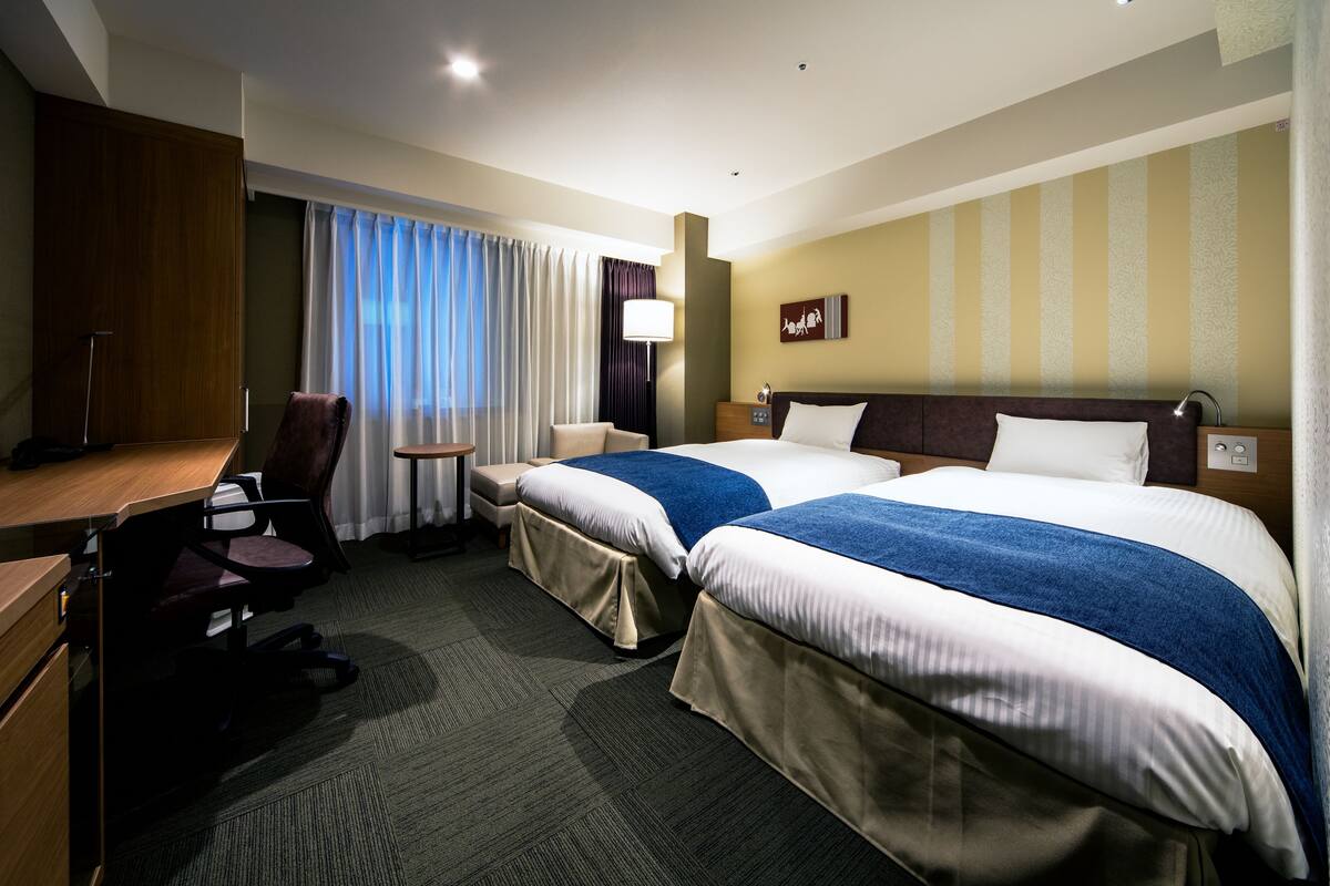 A hotel room with two beds, a desk, a chair, a lamp, and a window with curtains.