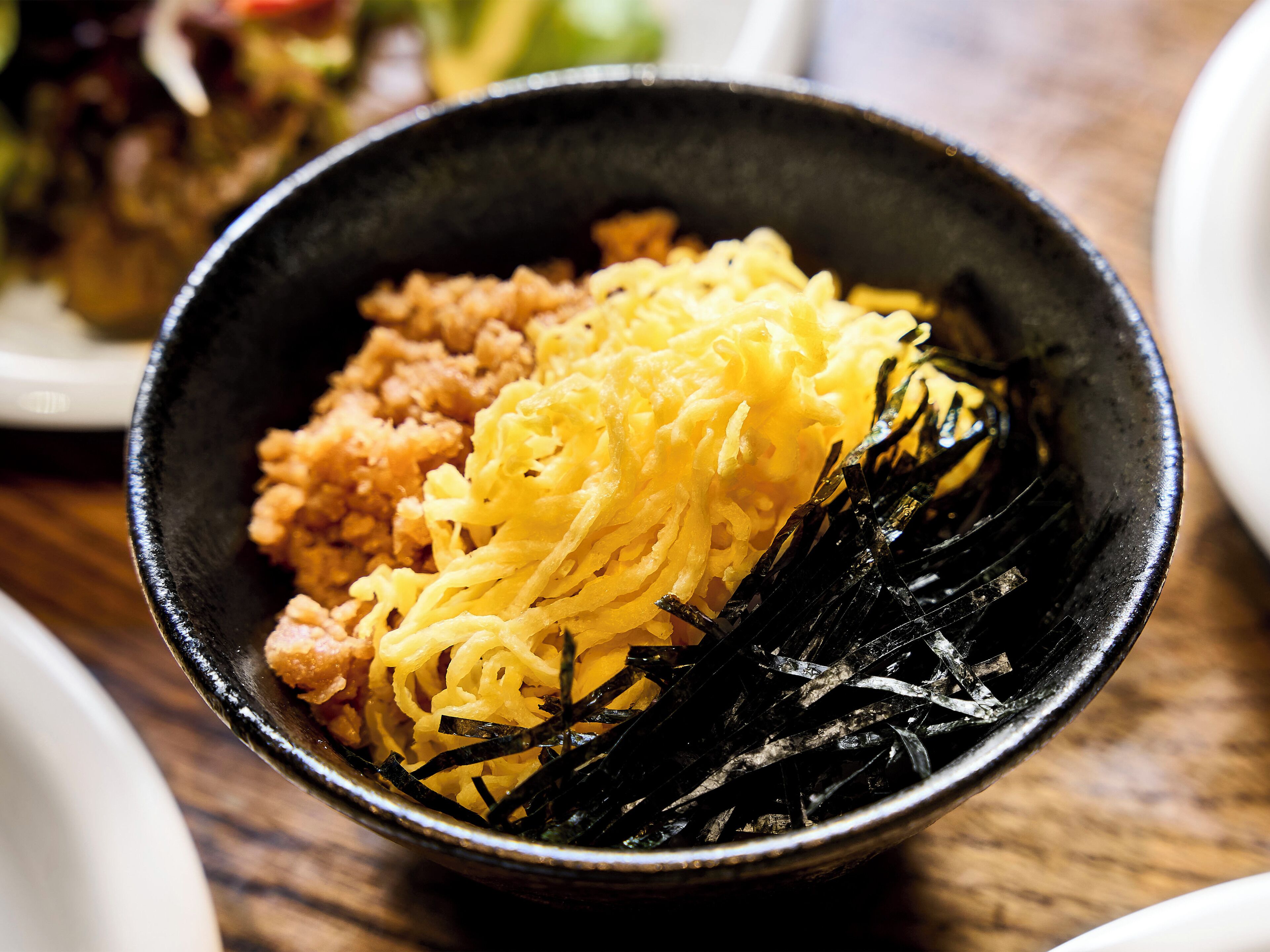 A bowl of food with noodles, a yellowish substance, and seaweed on a wooden table.