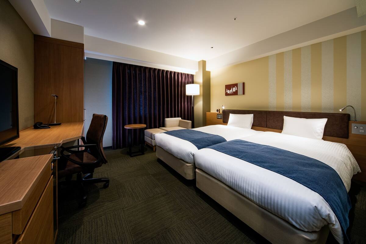 A hotel room with two beds, a desk, a chair, a lamp, and a nightstand.