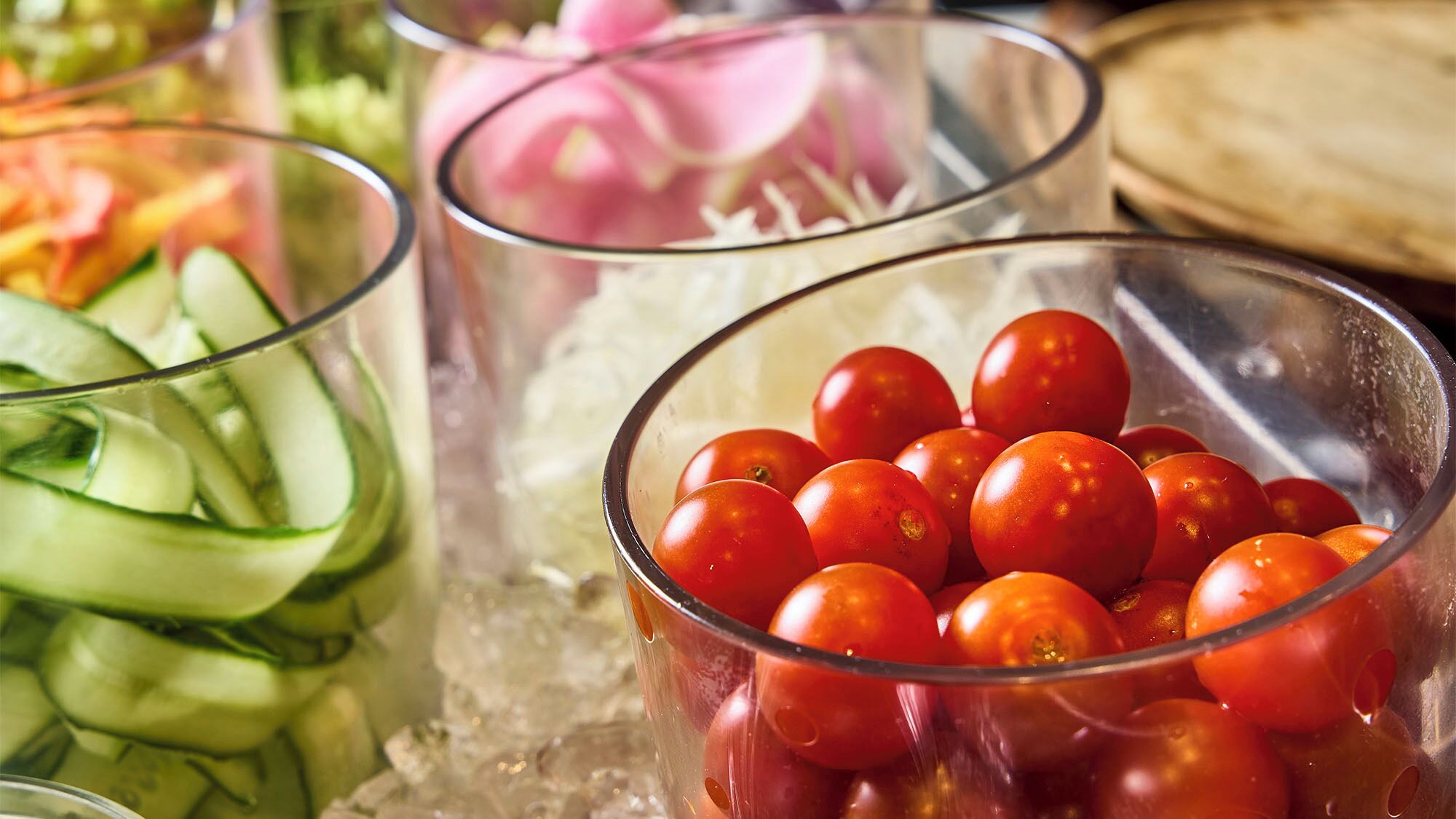 A glass bowl filled with cherry tomatoes on ice, a glass with sliced cucumbers, and a bowl of sliced onions.