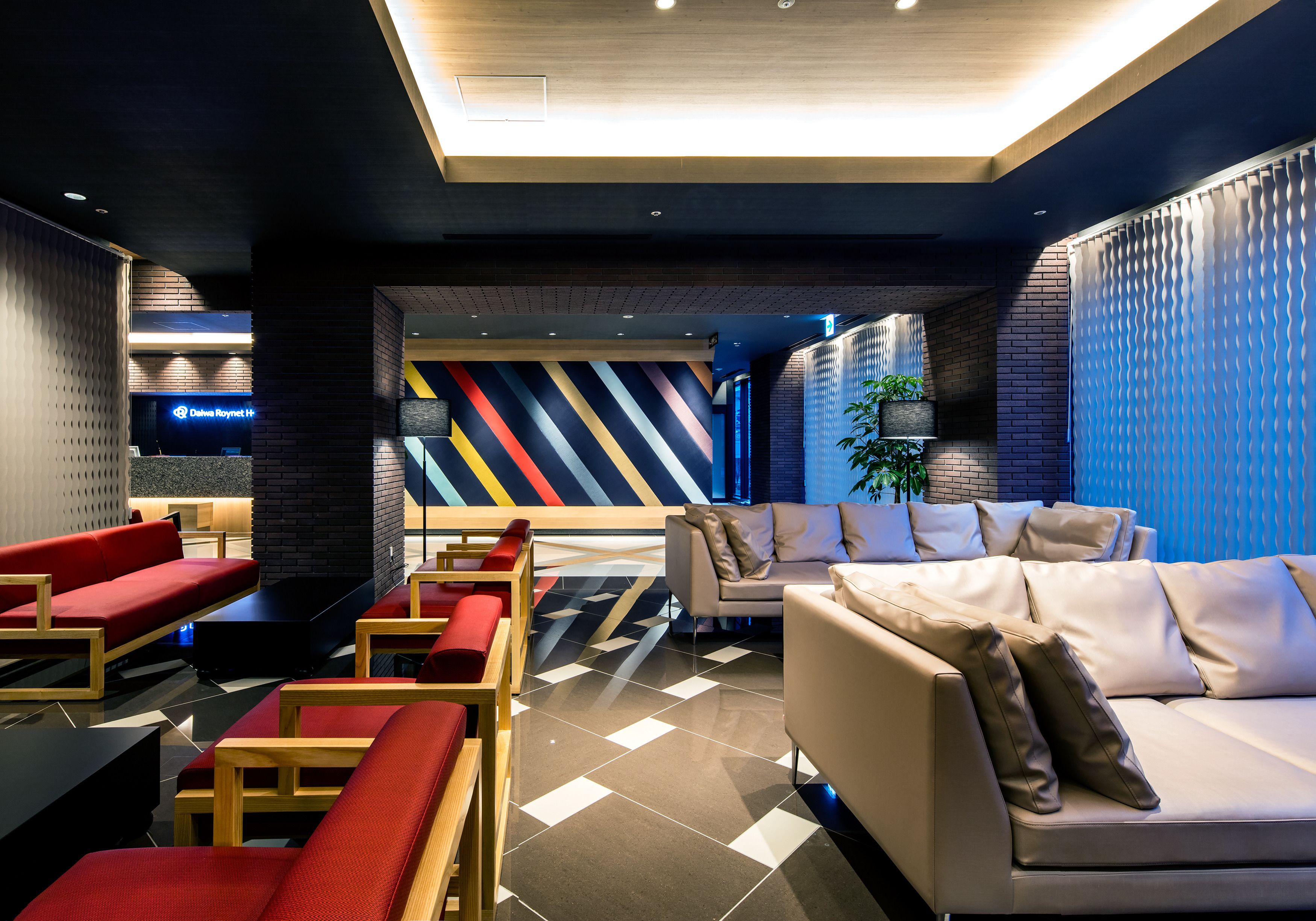 A modern lobby with a mix of seating areas, including red sofas and a beige sofa, and a patterned wall in the background.
