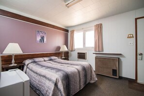 A hotel room with a bed, two bedside lamps, a window with curtains, a wall-mounted heater, and a small table with a lamp. - Wild Goose Motel and Campground (Kerrobert)