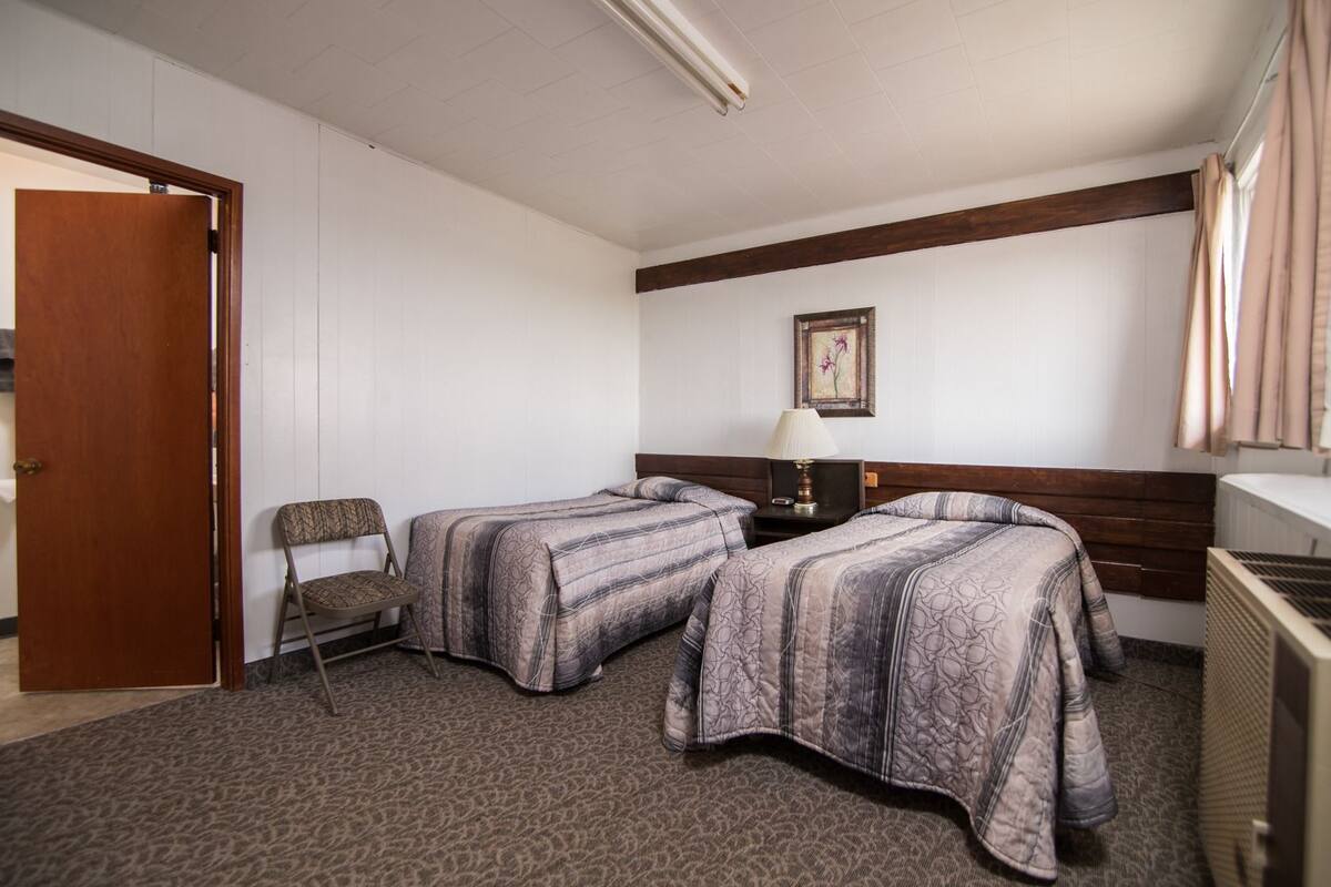 Quarto de hotel com duas camas, uma cadeira, um abajur e um quadro na parede.