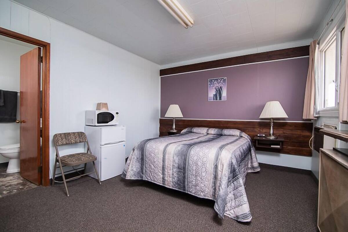 Quarto de hotel com uma cama, duas mesas de cabeceira com abajures, um micro-ondas, uma cadeira e um sanitário.