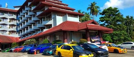 A multi-story hotel with a red roof and balconies, parking area with various cars, and tropical vegetation in the background.