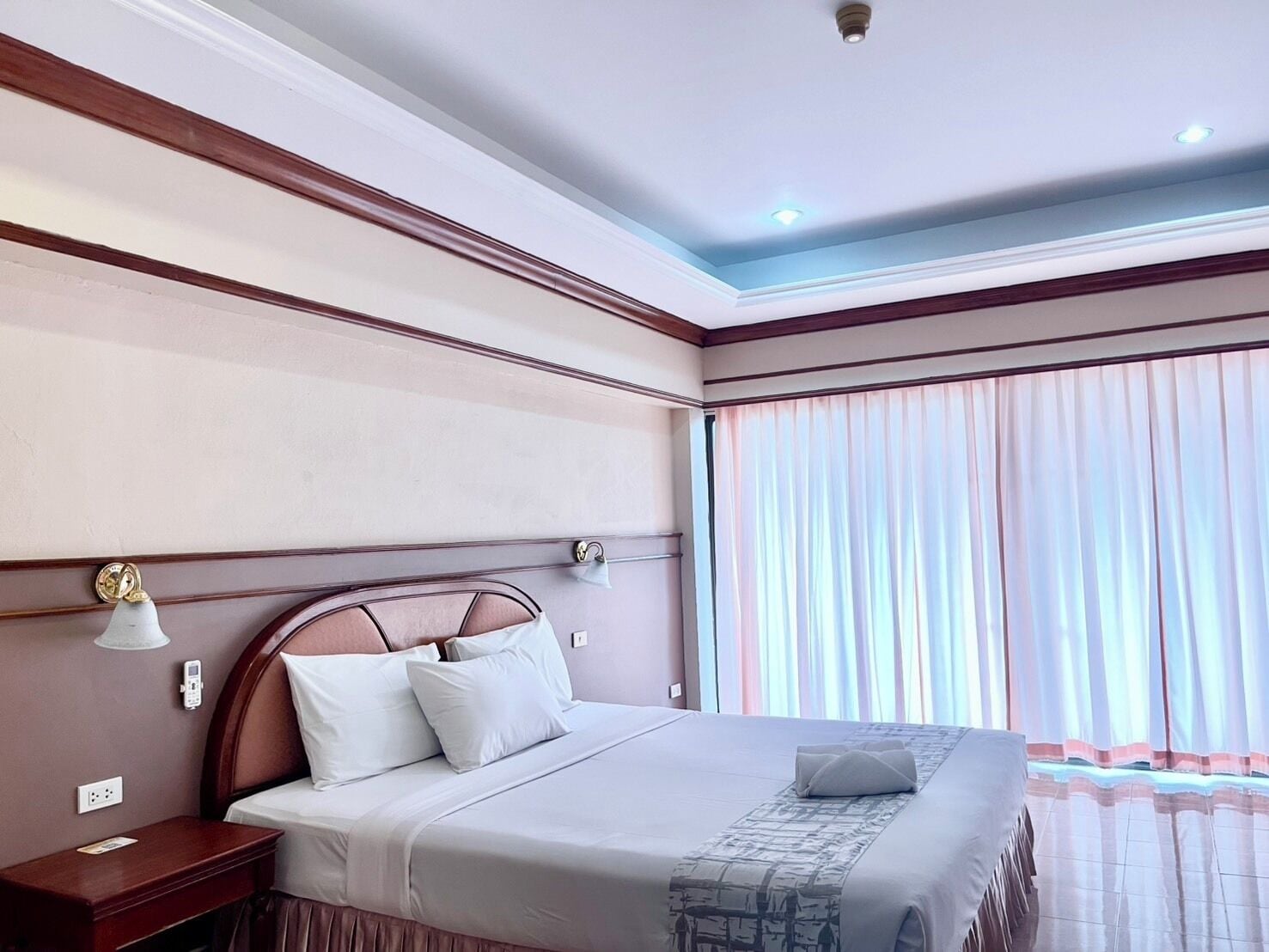 Habitación de hotel con una cama grande, una mesita de noche, una lámpara de pared y un ventanal con cortinas.