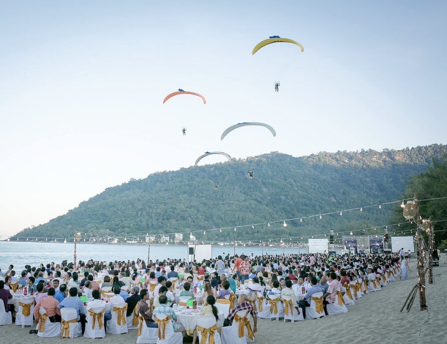 A beachside event with tables and chairs set up for an audience, and several parachutes in the sky.