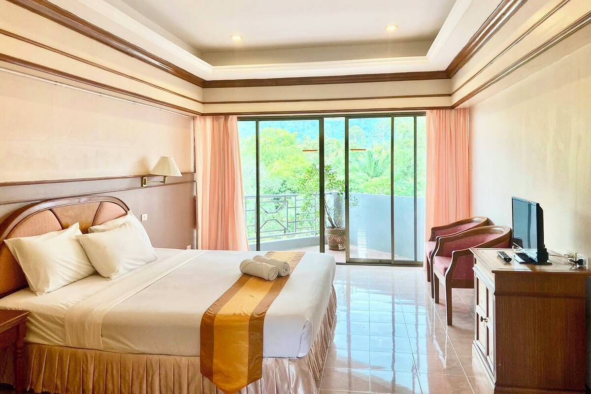 A hotel room with a large bed, a desk, a television, and a balcony with a view of greenery.