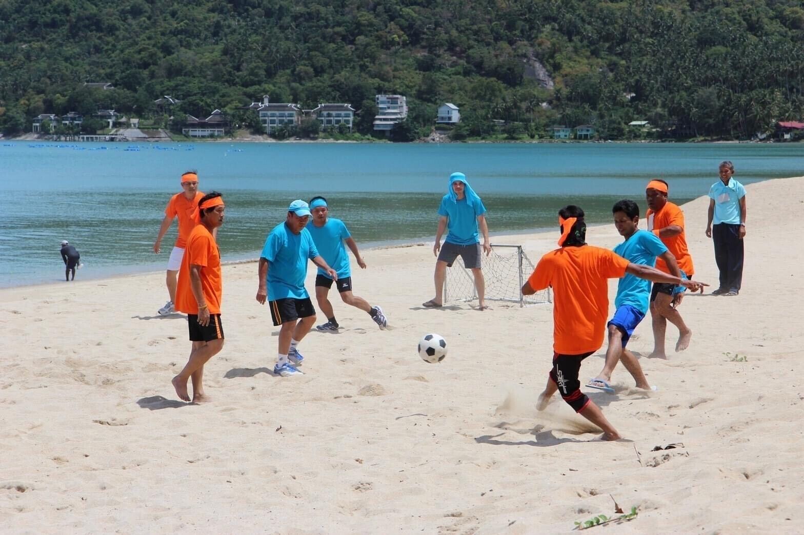 Un groupe de personnes joue au football sur une plage de sable, avec une étendue d’eau et des maisons en arrière-plan.