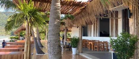 A wooden deck with thatched roof seating, a dining area with wooden tables and chairs, and a large potted plant.