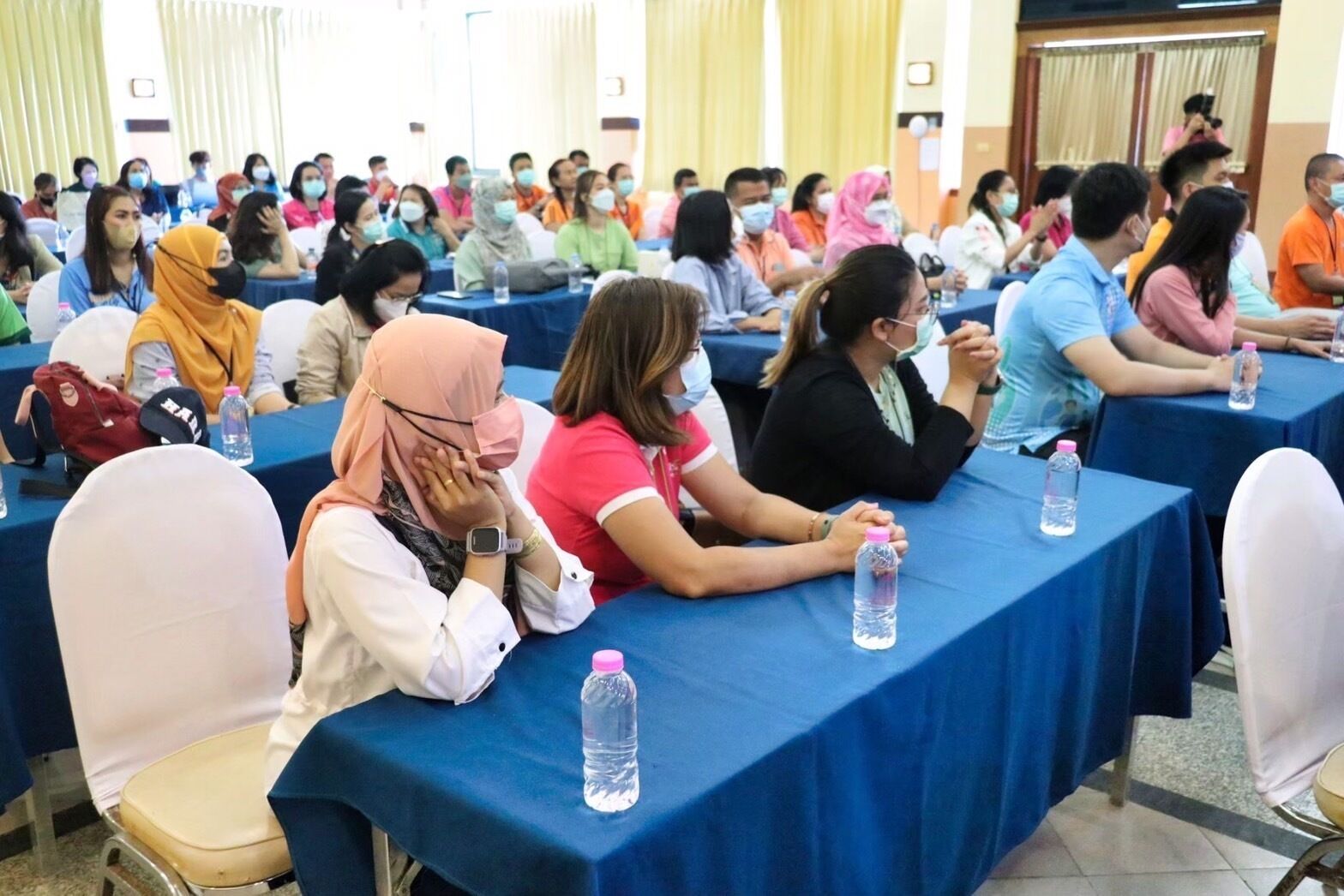 A conference room with rows of tables, attendees seated, and water bottles on tables.
