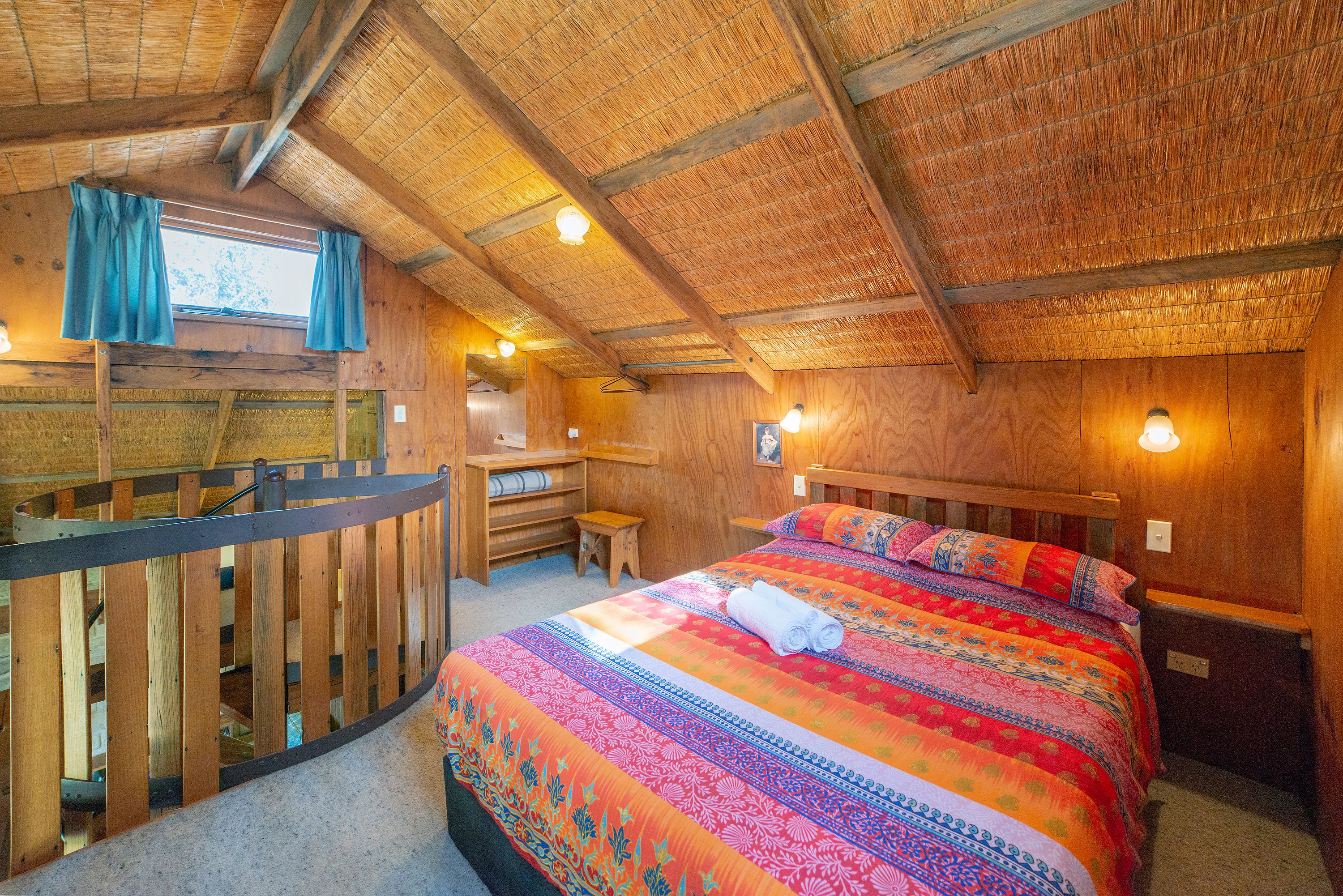 A cozy room with a wooden ceiling, a bed with a colorful blanket, a desk, and a small stool.
