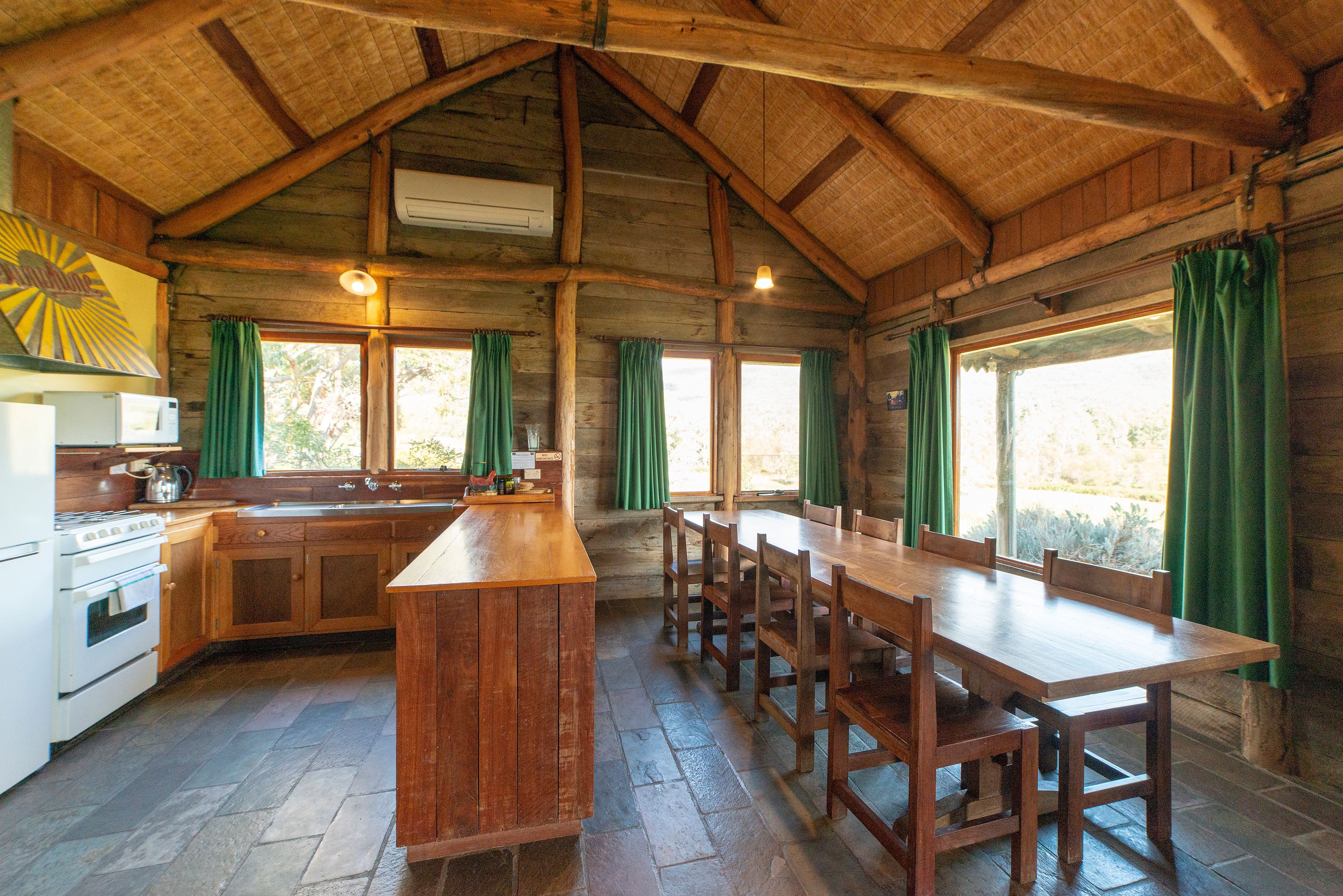 Un chalet rustique avec un intérieur en bois, une cuisine dotée d’un îlot central et de grandes fenêtres ornées de rideaux verts.