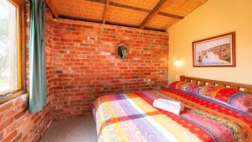 Una habitación con pared de ladrillo, techo de madera, cama con colcha colorida, un cuadro enmarcado en la pared y ventana con cortinas.