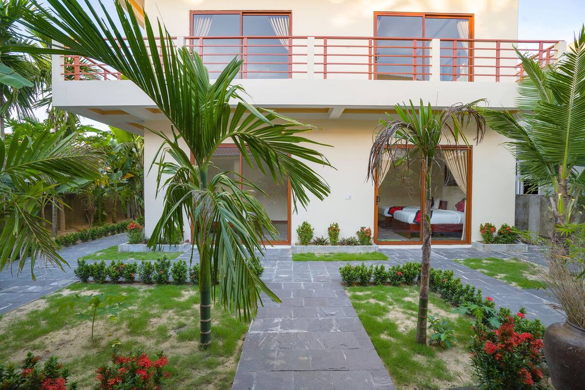 A modern two-story building with a balcony, a pathway leading to the entrance, and a garden with palm trees and red flowers.