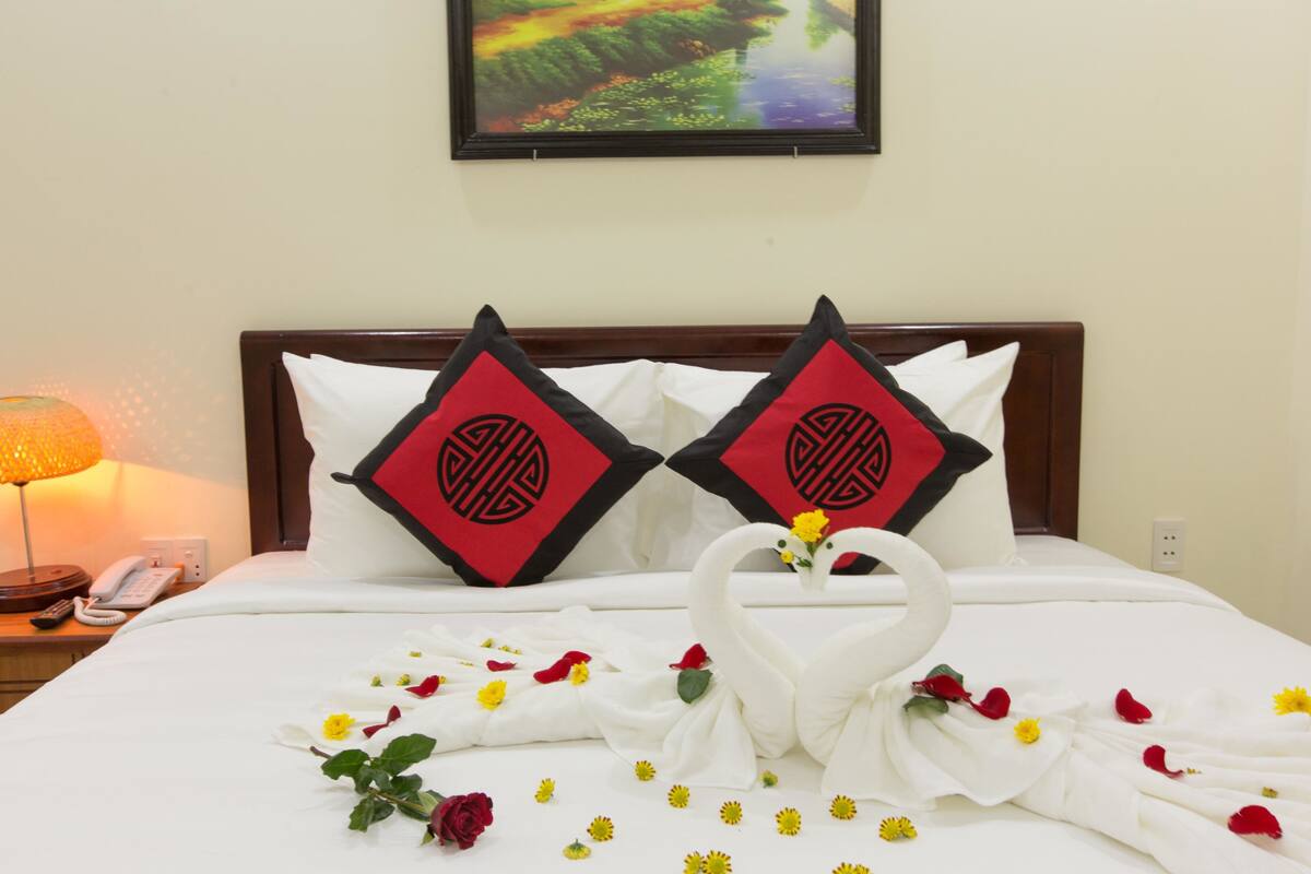 A neatly made bed with a swan-shaped towel arrangement, two pillows with a red and black design, and a framed picture on the wall.