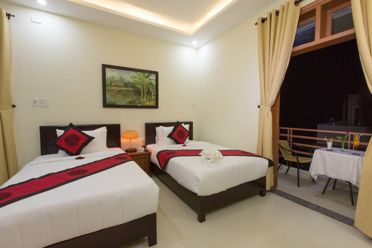 A hotel room with two beds, a nightstand with a lamp, a painting on the wall, and a balcony with a table and chairs.