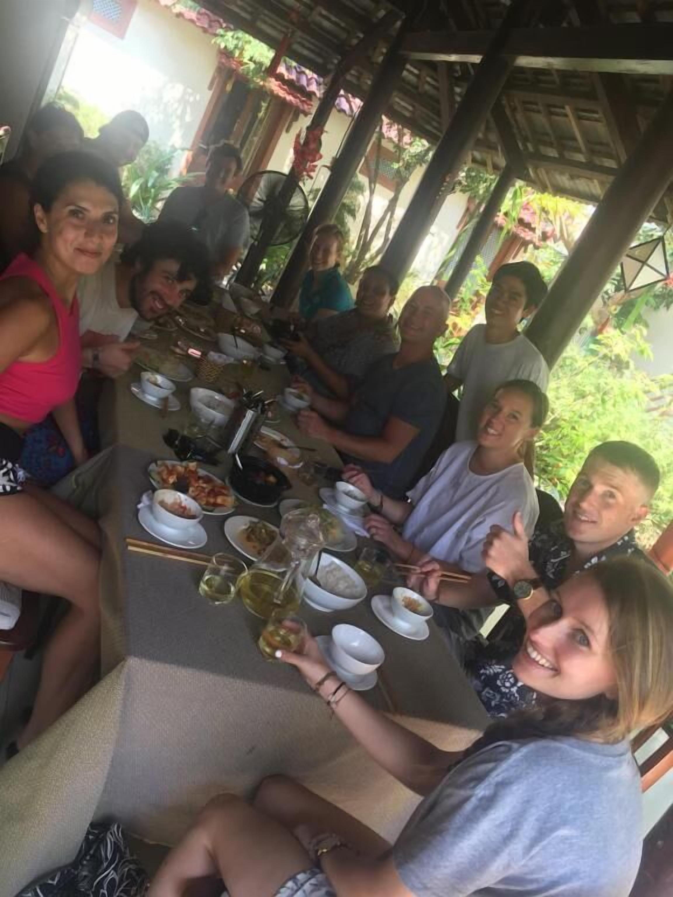 A group of people seated around a table with various dishes and drinks, under a covered outdoor area with a wooden structure.