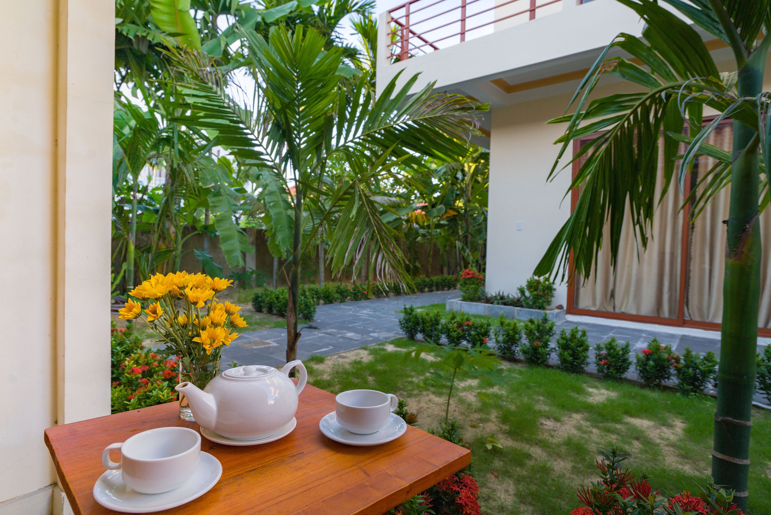 A wooden table with a teapot and cups, a vase of yellow flowers, and a view of a garden with a pathway and greenery.