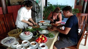 A dining table with various dishes and condiments, a fan, and a refrigerator in the background. - Herbal Tea Homestay (Da Nang)