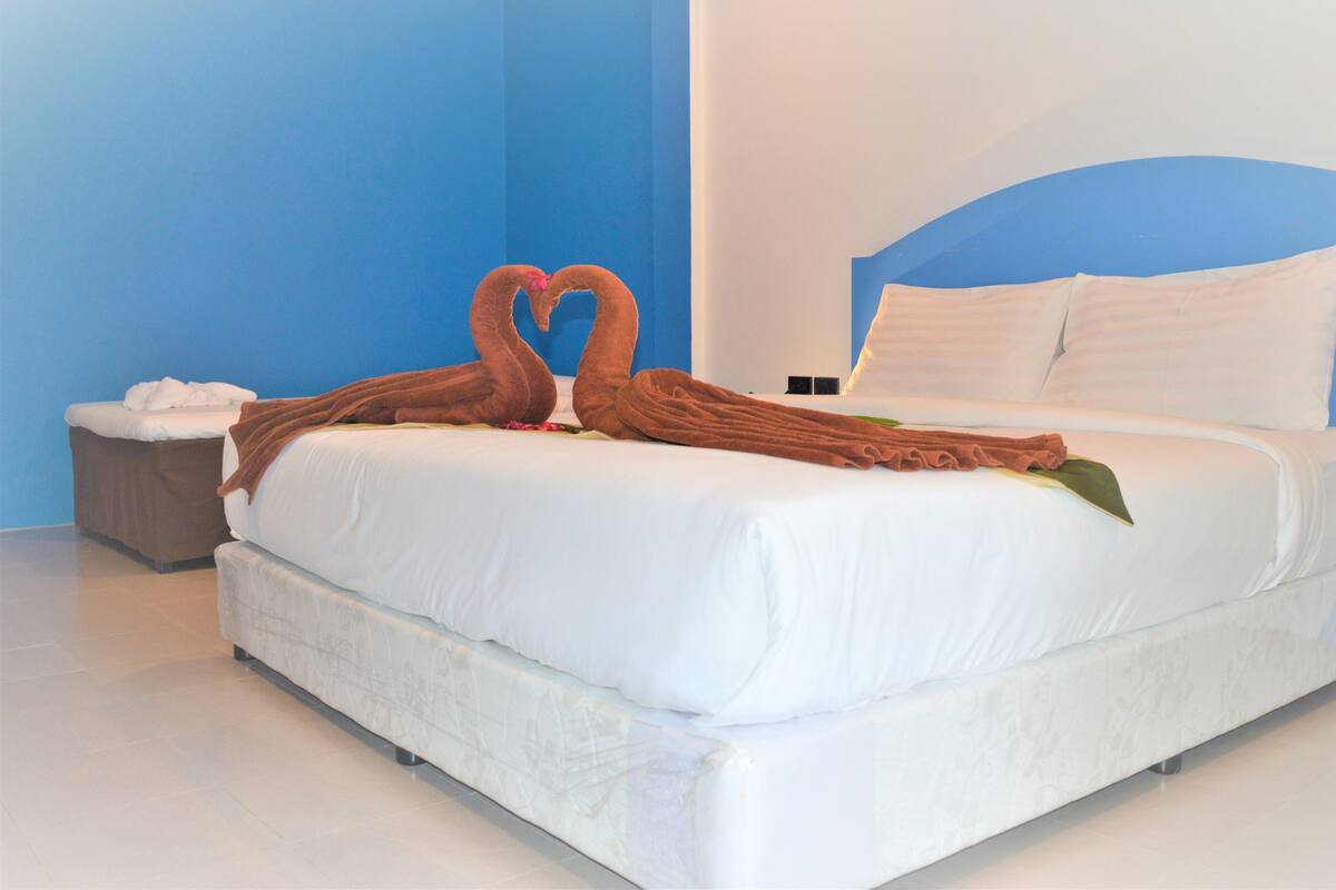 A neatly made bed with white linens, a brown towel arranged in a heart shape, and a small bedside table with a tissue box.
