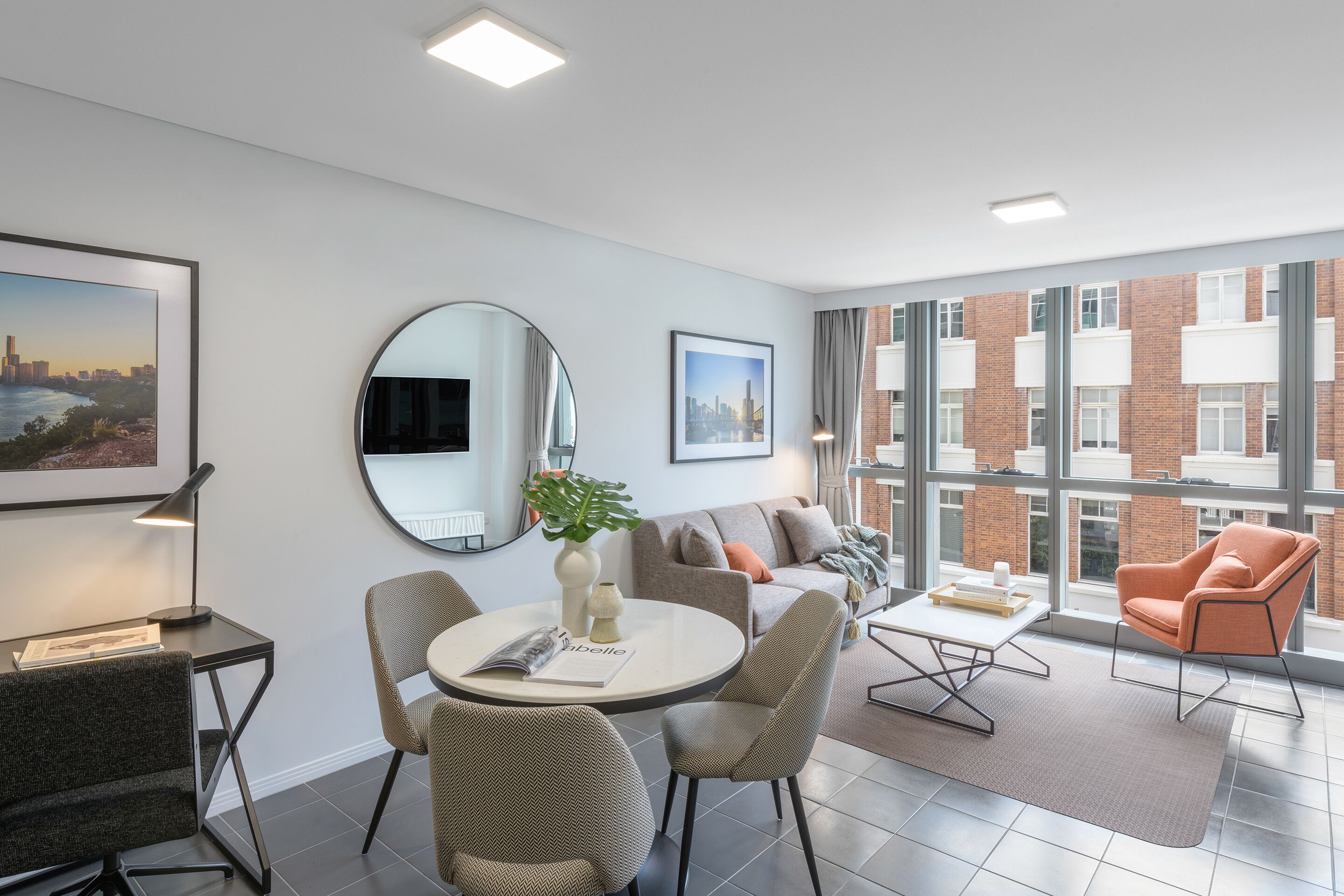A modern hotel room with a dining area, a round table with chairs, a sofa, a large window with a city view, and a mirror on the wall.