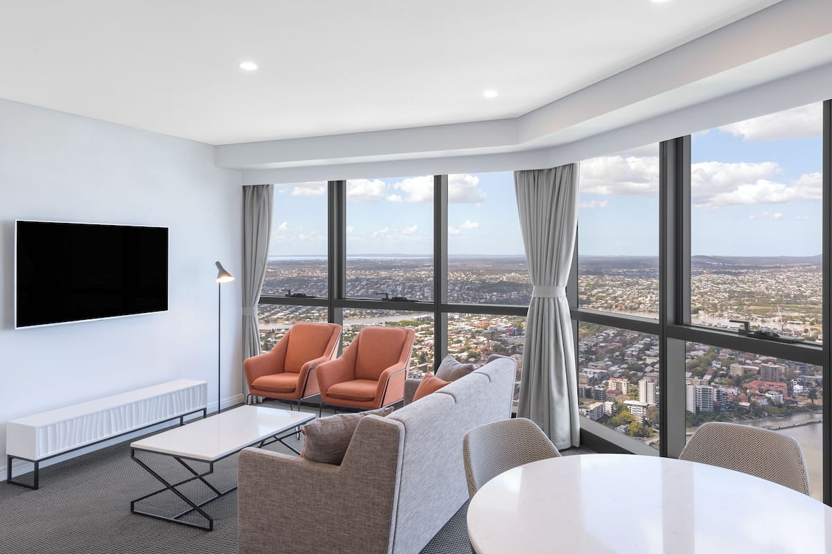 Habitación de hotel moderna con un amplio ventanal que ofrece vistas a la ciudad, un televisor de pantalla plana, un sofá y una mesa de comedor.