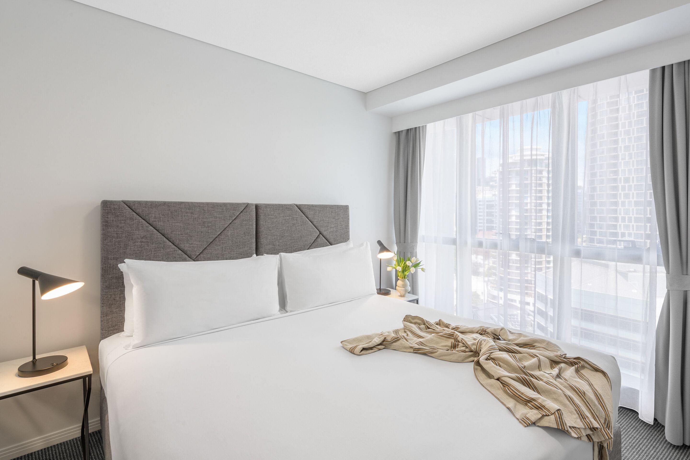A hotel room with a neatly made bed, a bedside lamp, and a large window with curtains.